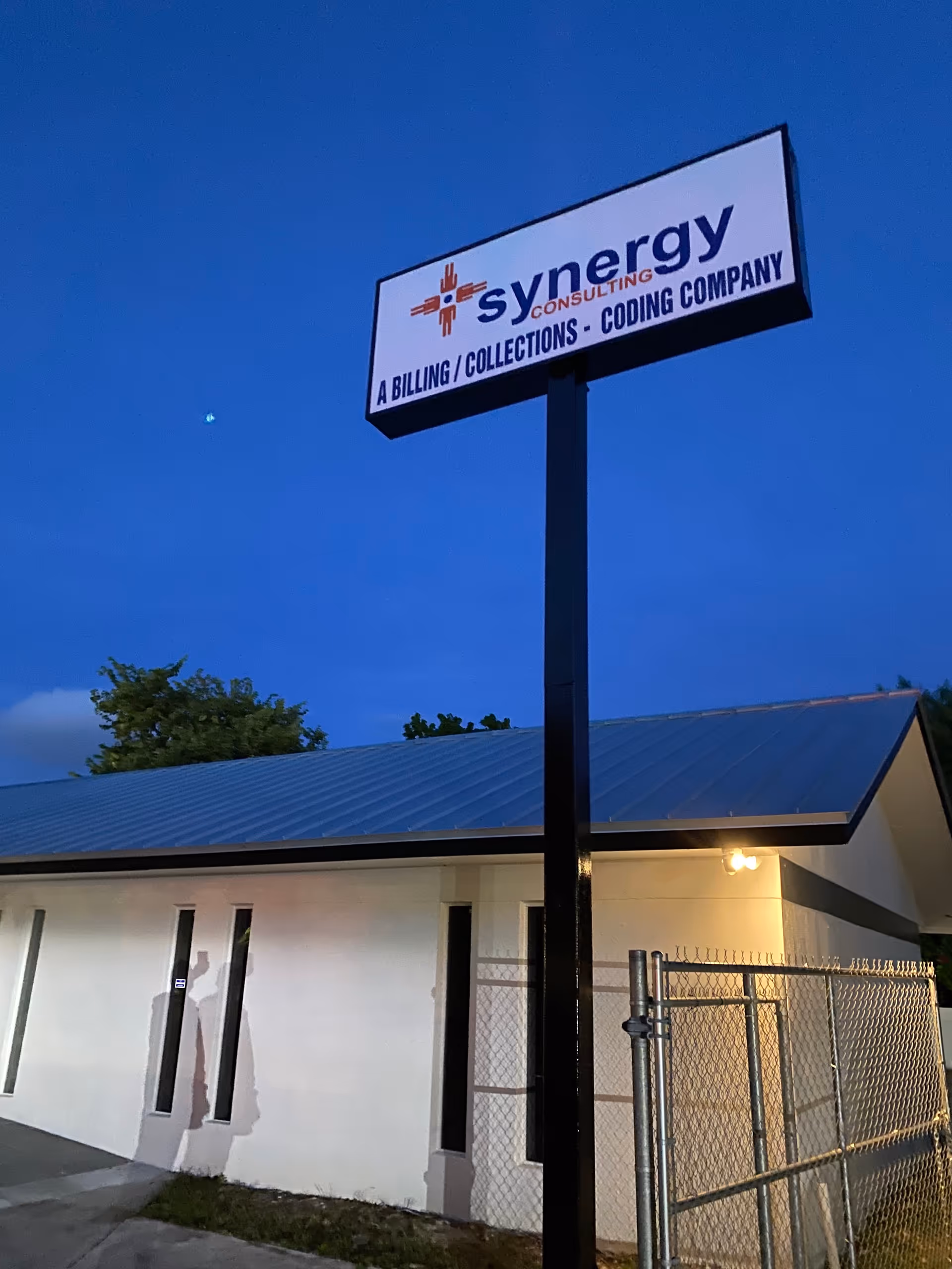 Exterior view of a white building with a tall Synergy sign against a blue evening sky.