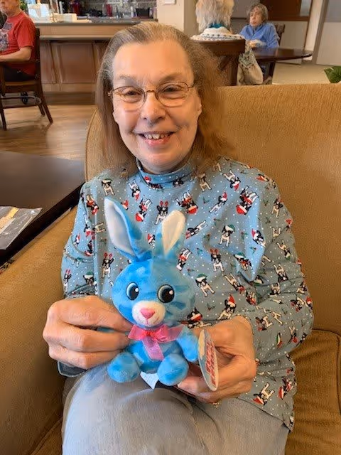 An elderly woman with glasses sitting on a beige couch, smiling and holding a blue stuffed bunny with white ears and a pink bow. She is wearing a light blue shirt with a pattern of small dogs in red and white outfits. In the background, other elderly people are seated in a common area with wooden flooring and furniture.