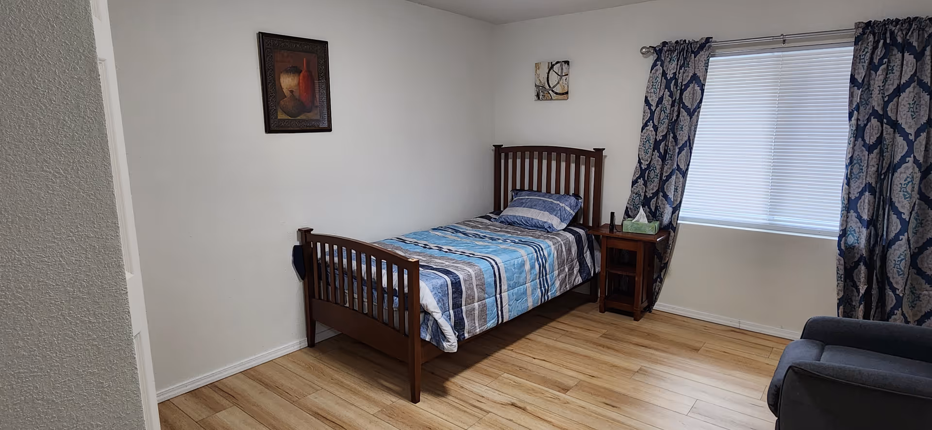A single wooden bed with blue striped bedding sits in a tidy room with curtains.