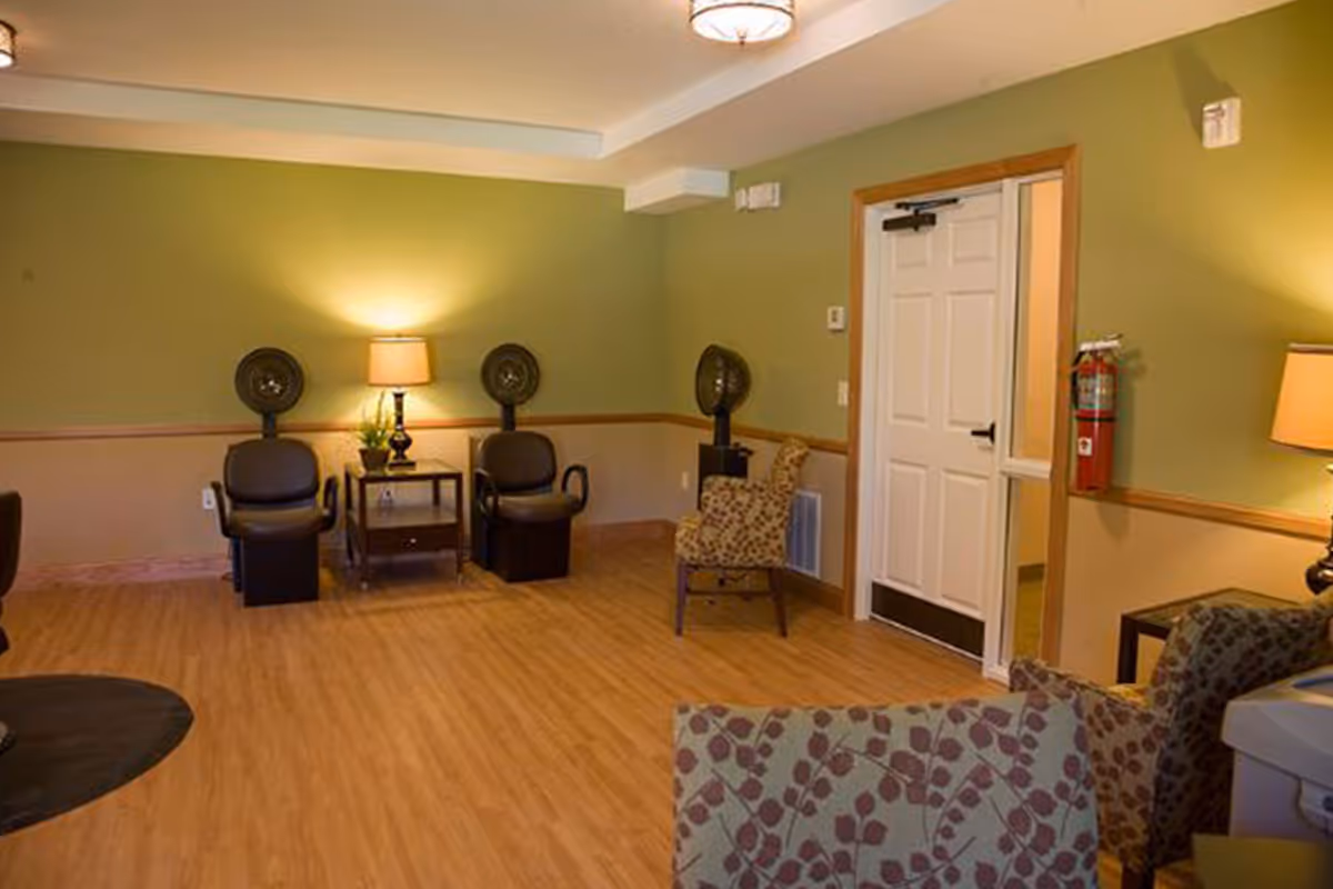 Well-lit common lounge with seating, side tables, lamps, wall-mounted hair-dryer stations, and a doorway.