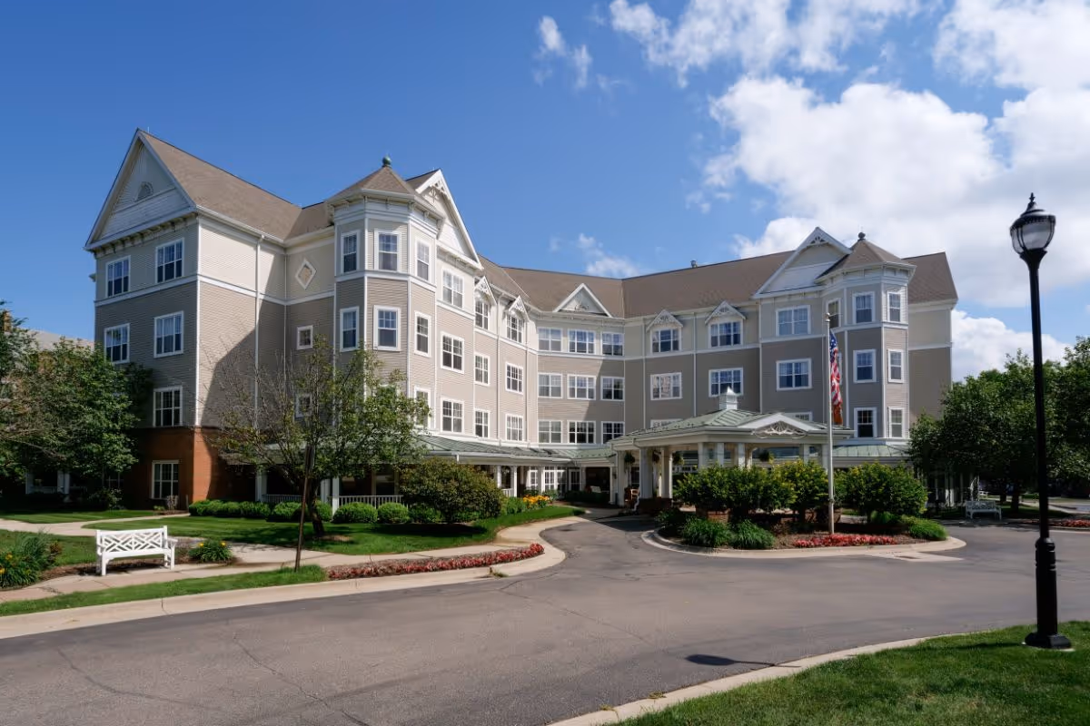 Exterior view of a large, multi-story assisted living facility building with beige siding and white trim under a partly cloudy blue sky. The building has multiple windows, a covered entrance with a circular driveway, landscaped greenery, a white bench, and a street lamp.