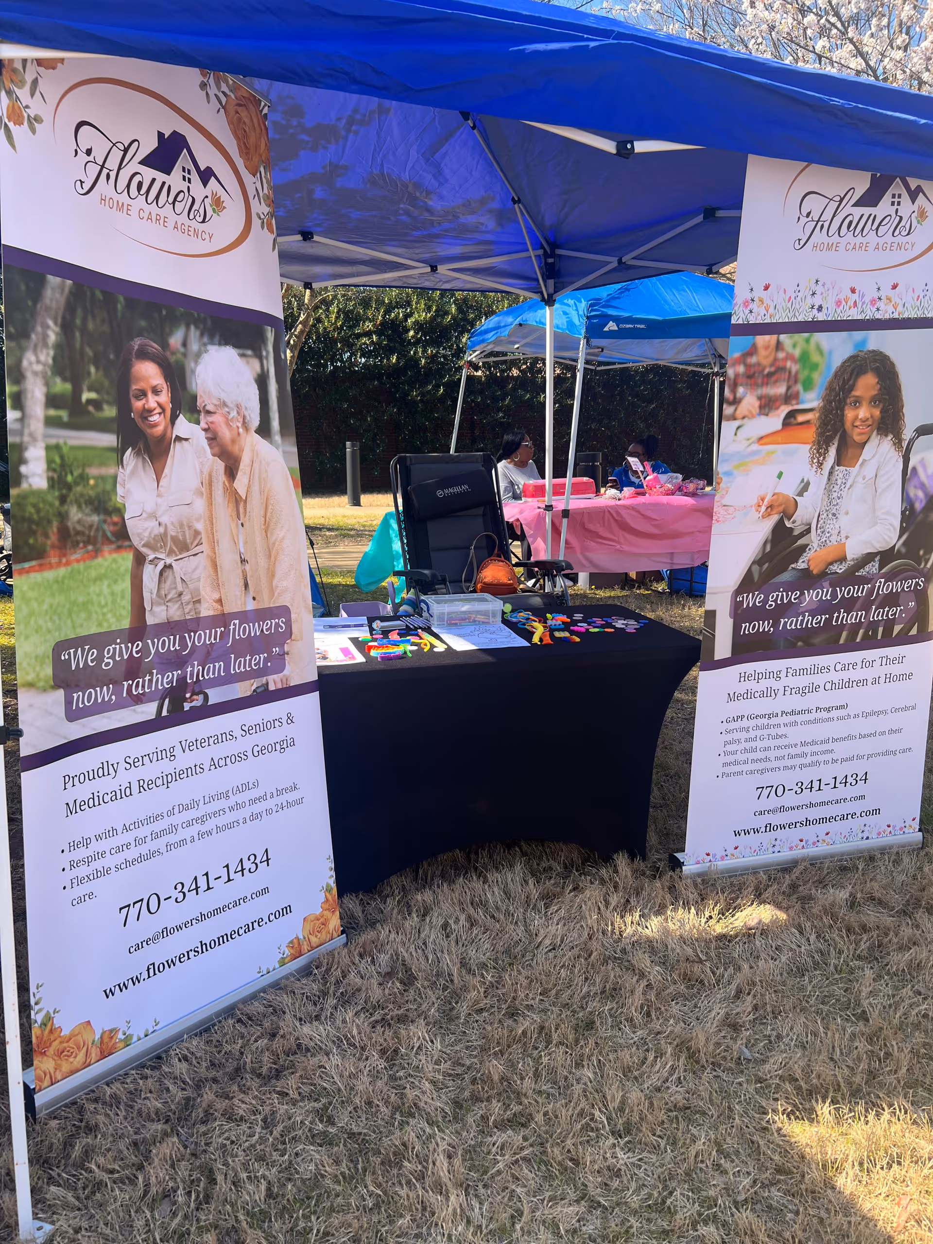 Outdoor Flowers Home Care Agency booth with banners, tables, and tents on grass.
