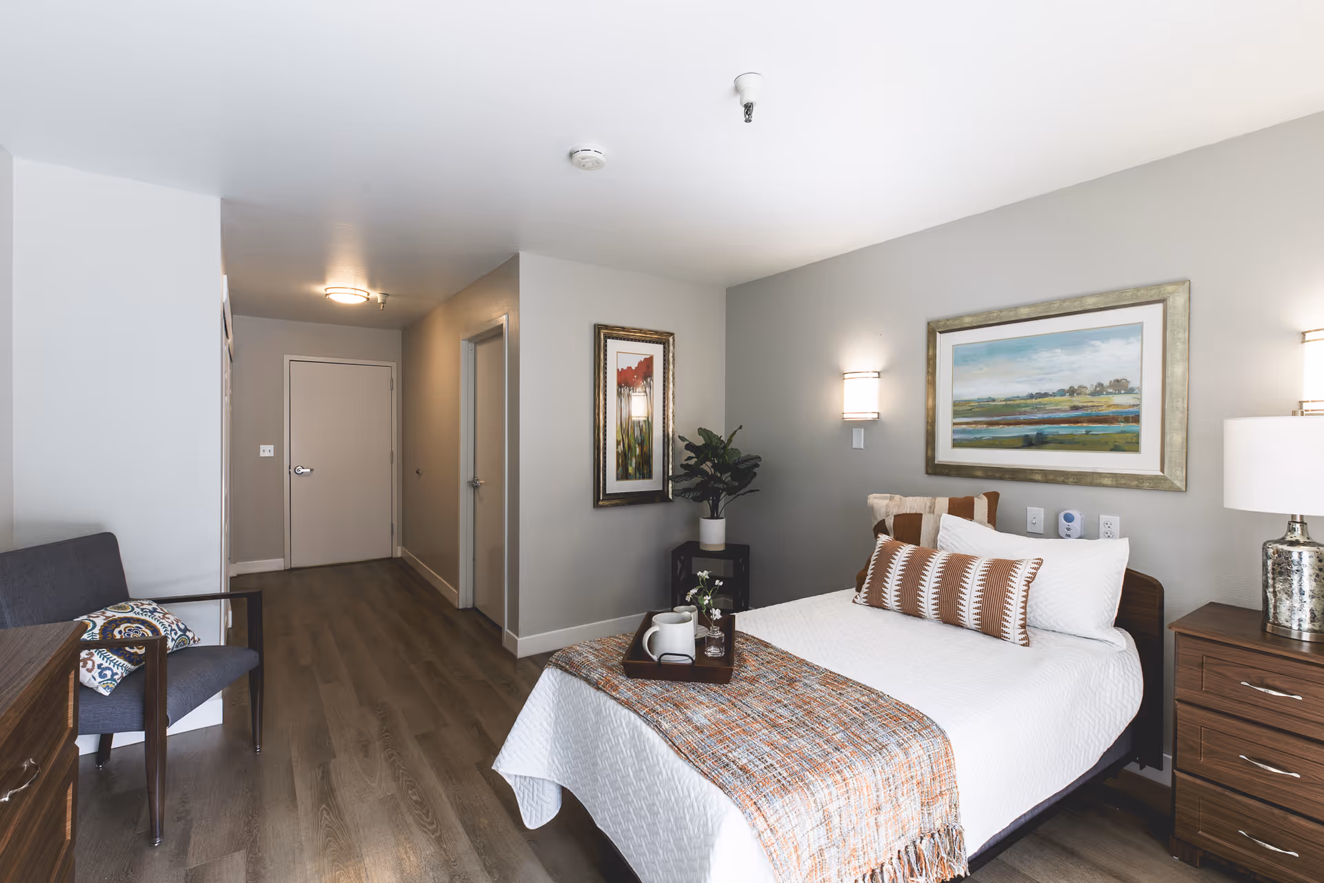 A furnished senior living bedroom with bed, nightstand, artwork, and seating.