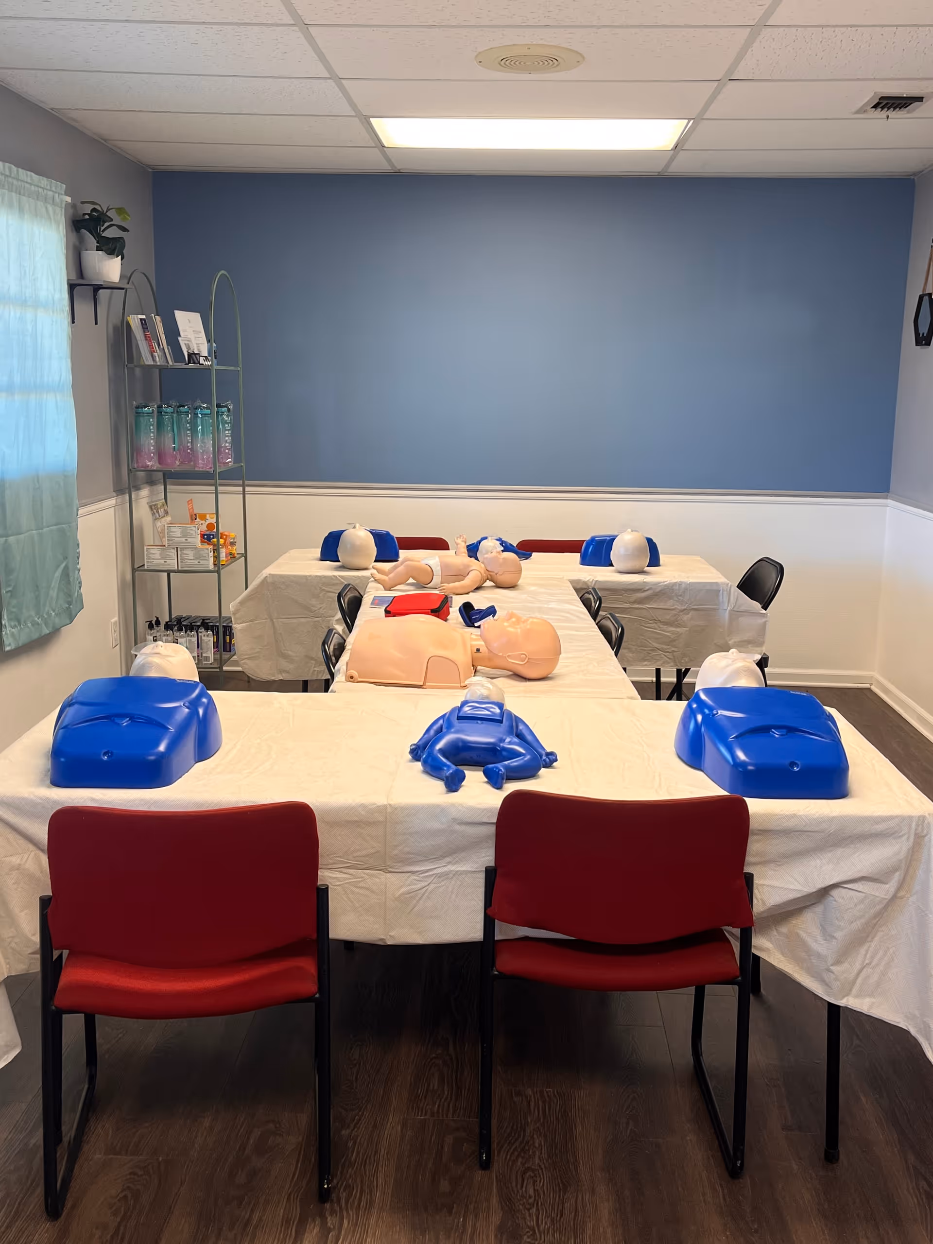 CPR training room with mannequins, tables, red chairs, and a blue wall.