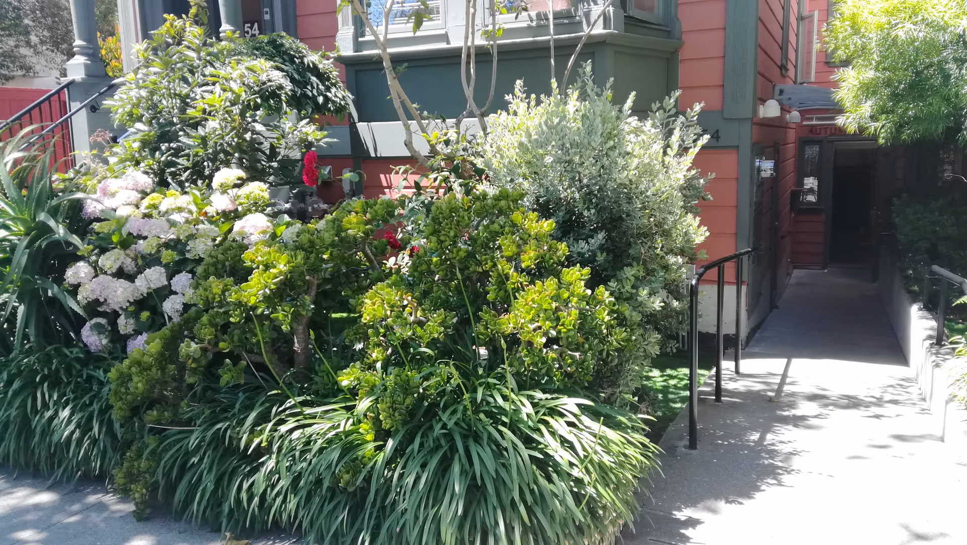 Entrance to a residential building named Autumn Glow with a pathway and handrails leading to the door, surrounded by lush green bushes and flowering plants.
