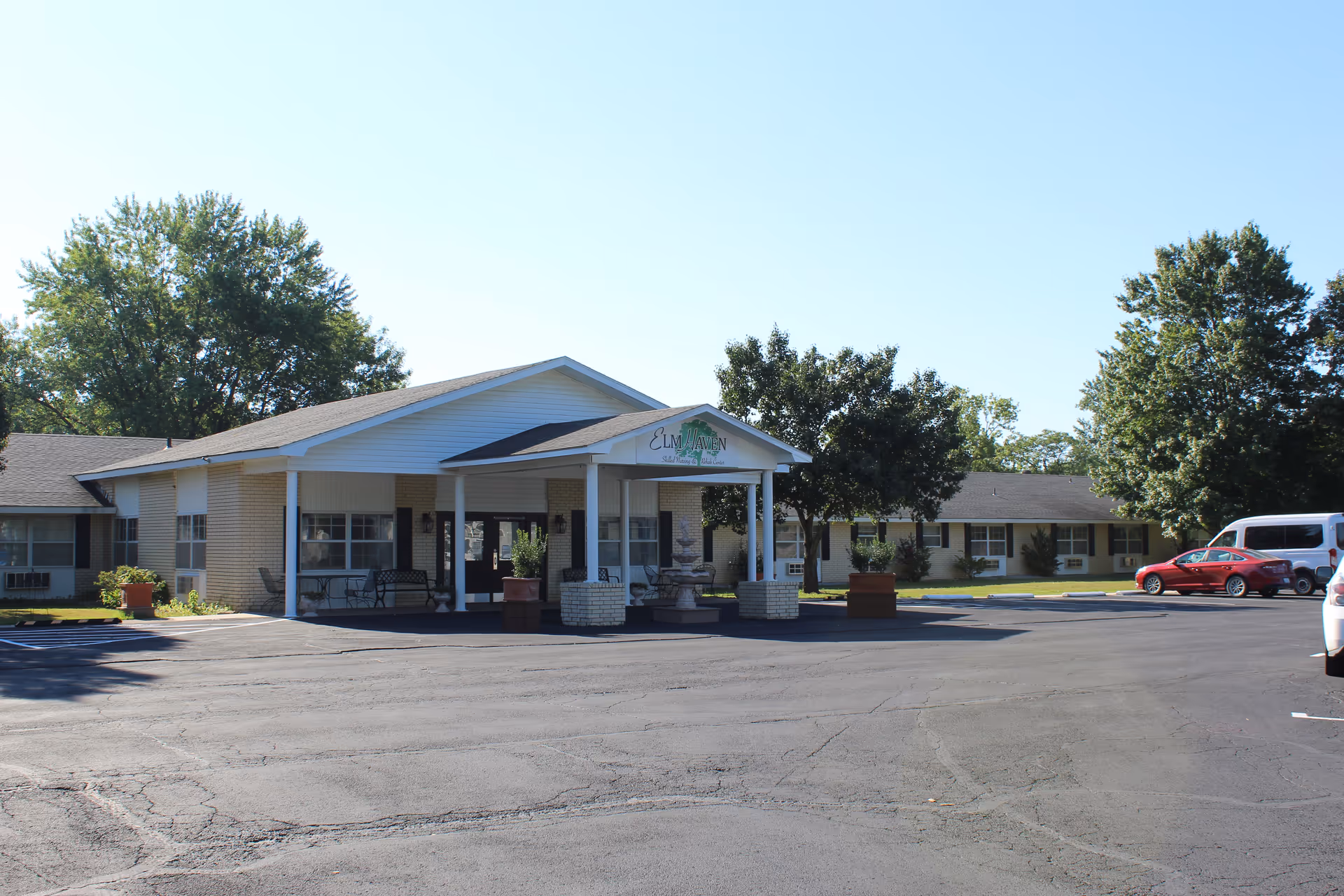 Exterior view of Elmhaven facility showing a single-story building with a covered entrance supported by white columns. There are benches and potted plants near the entrance, a parking lot with several cars, and trees surrounding the building under a clear blue sky.