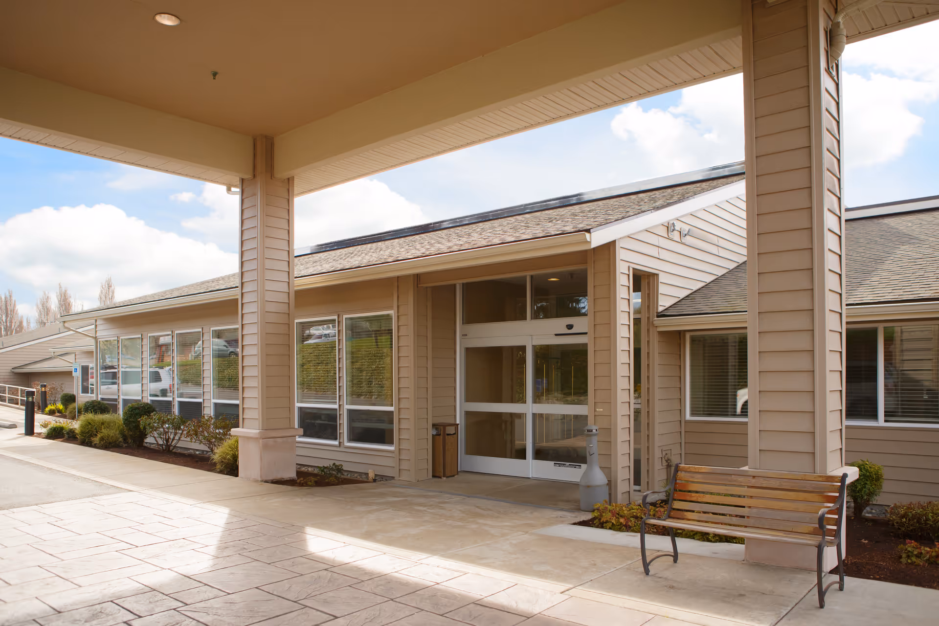 Entrance of a single-story building with beige siding and large windows, featuring a covered walkway supported by columns. A wooden bench is placed near one of the columns, and there are some small bushes and plants along the building. The sky is partly cloudy.