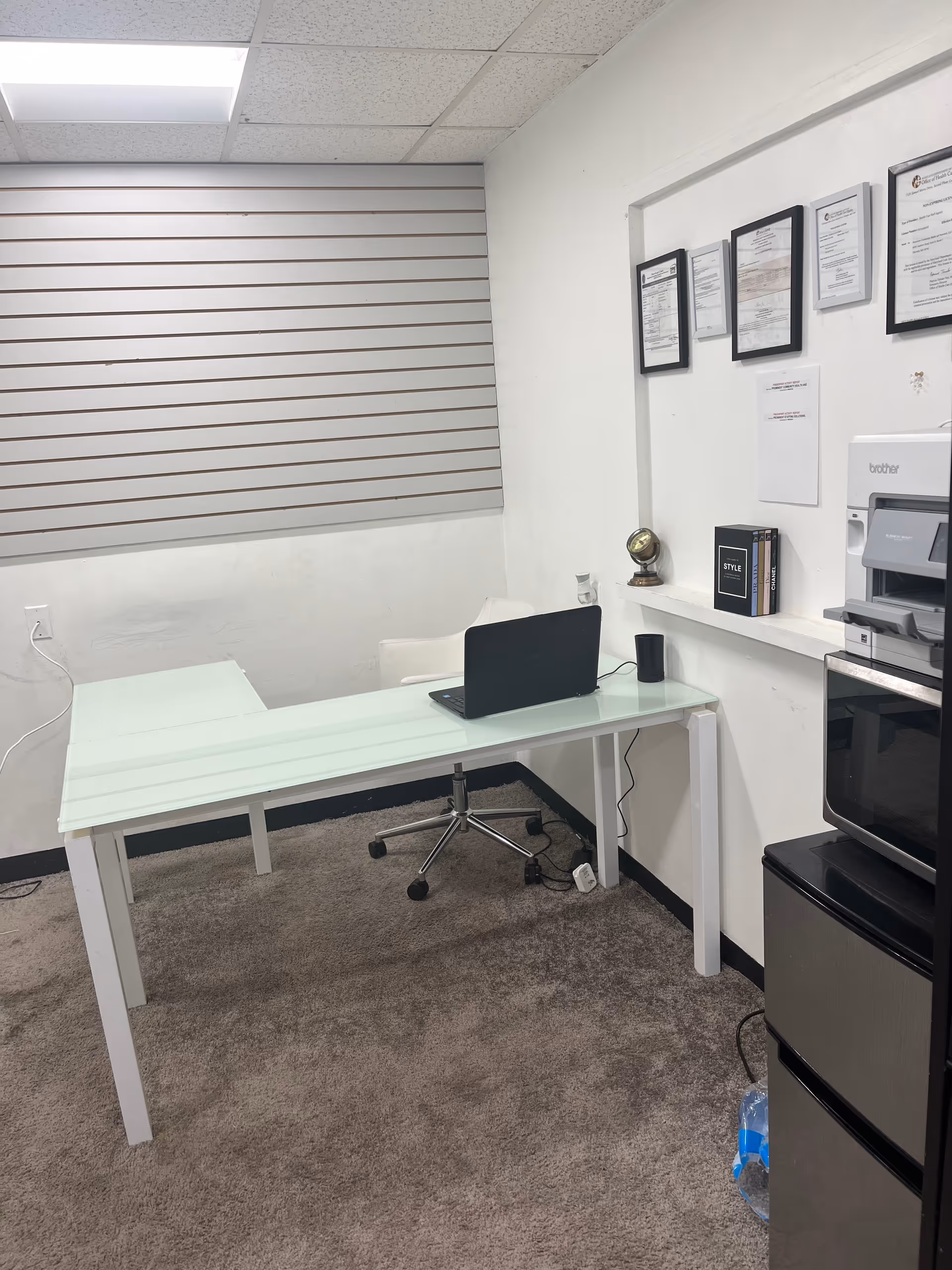 Office workspace with a glass desk, laptop, chair, and filing area in a white room.