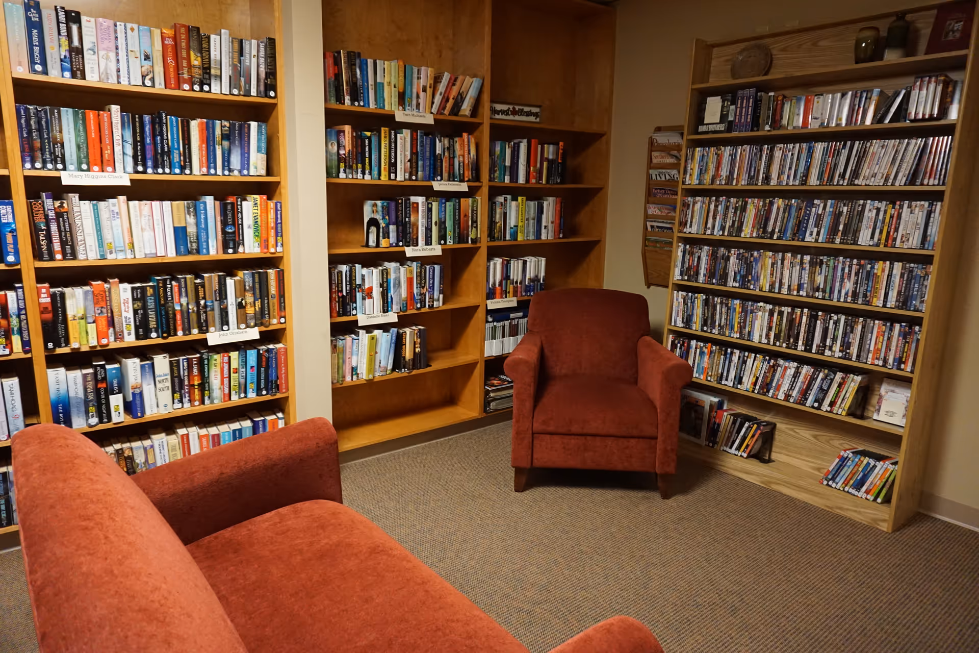 Cozy reading room with wooden bookshelves filled with books and DVDs, featuring a red upholstered armchair and a matching red sofa on a carpeted floor.