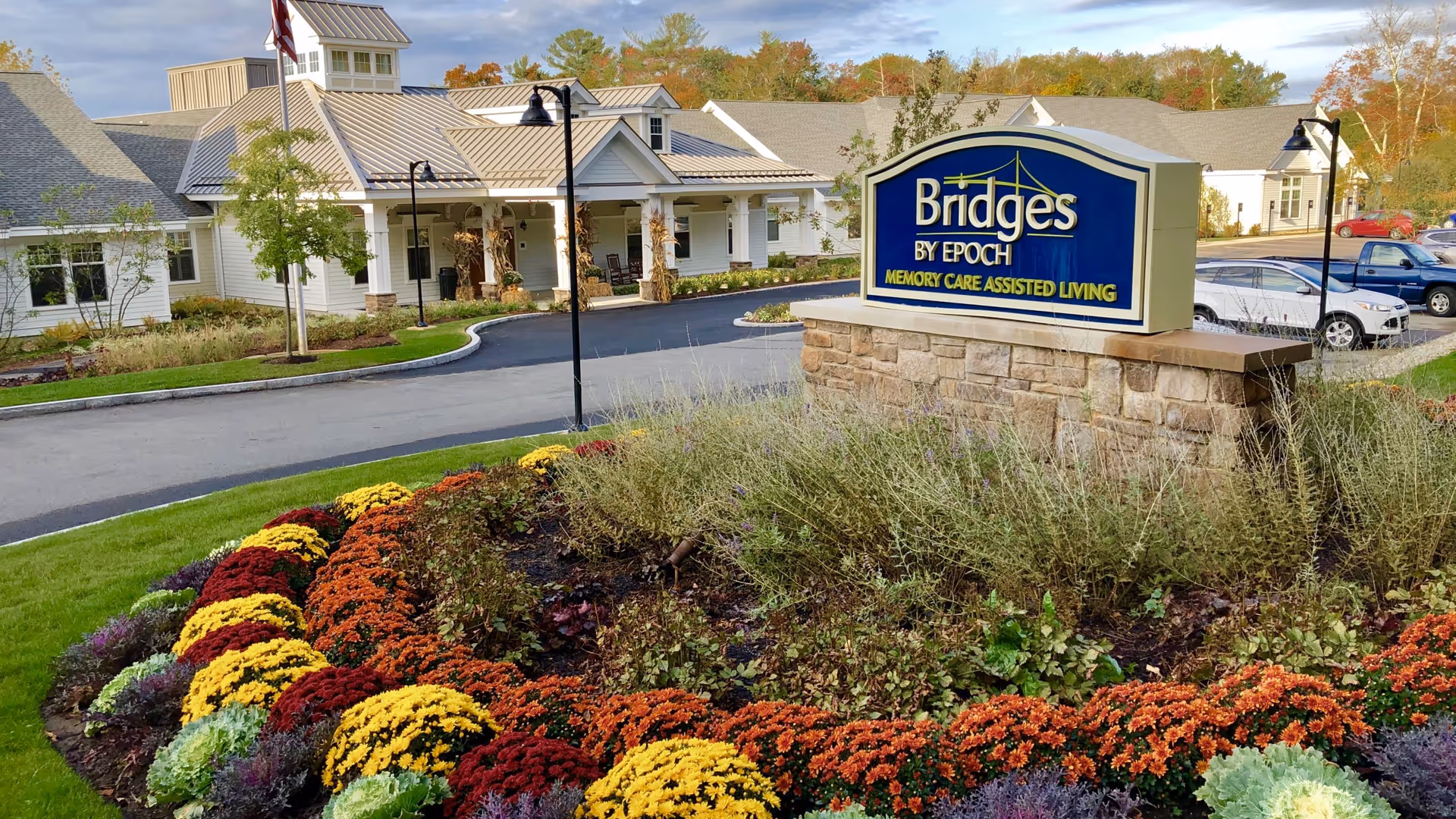 Exterior view of Bridges by EPOCH at Pembroke, a memory care assisted living facility, showing the building with a covered entrance, landscaped garden with colorful flowers, a stone sign with the facility name, and a parking area with cars.