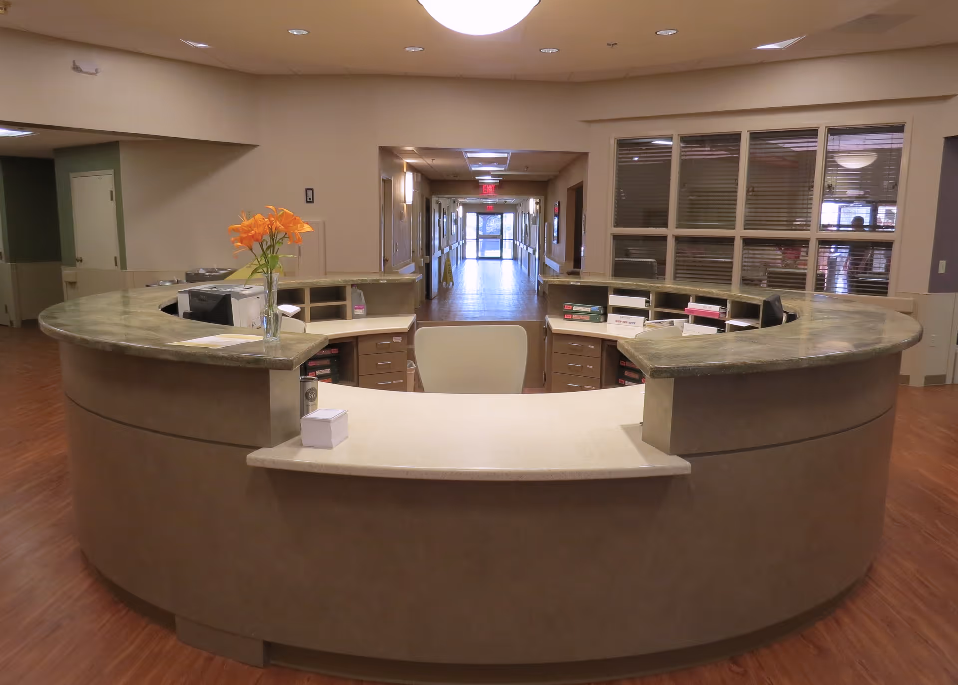 A curved reception desk in a nursing and rehabilitation center with a vase of orange flowers on the counter. Behind the desk are office supplies, binders, and a chair. A hallway extends into the background with doors and windows visible.