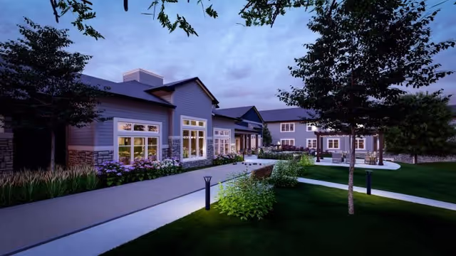Exterior view of a senior living facility building at dusk with well-lit windows, landscaped greenery, trees, and a paved walkway leading to the entrance.