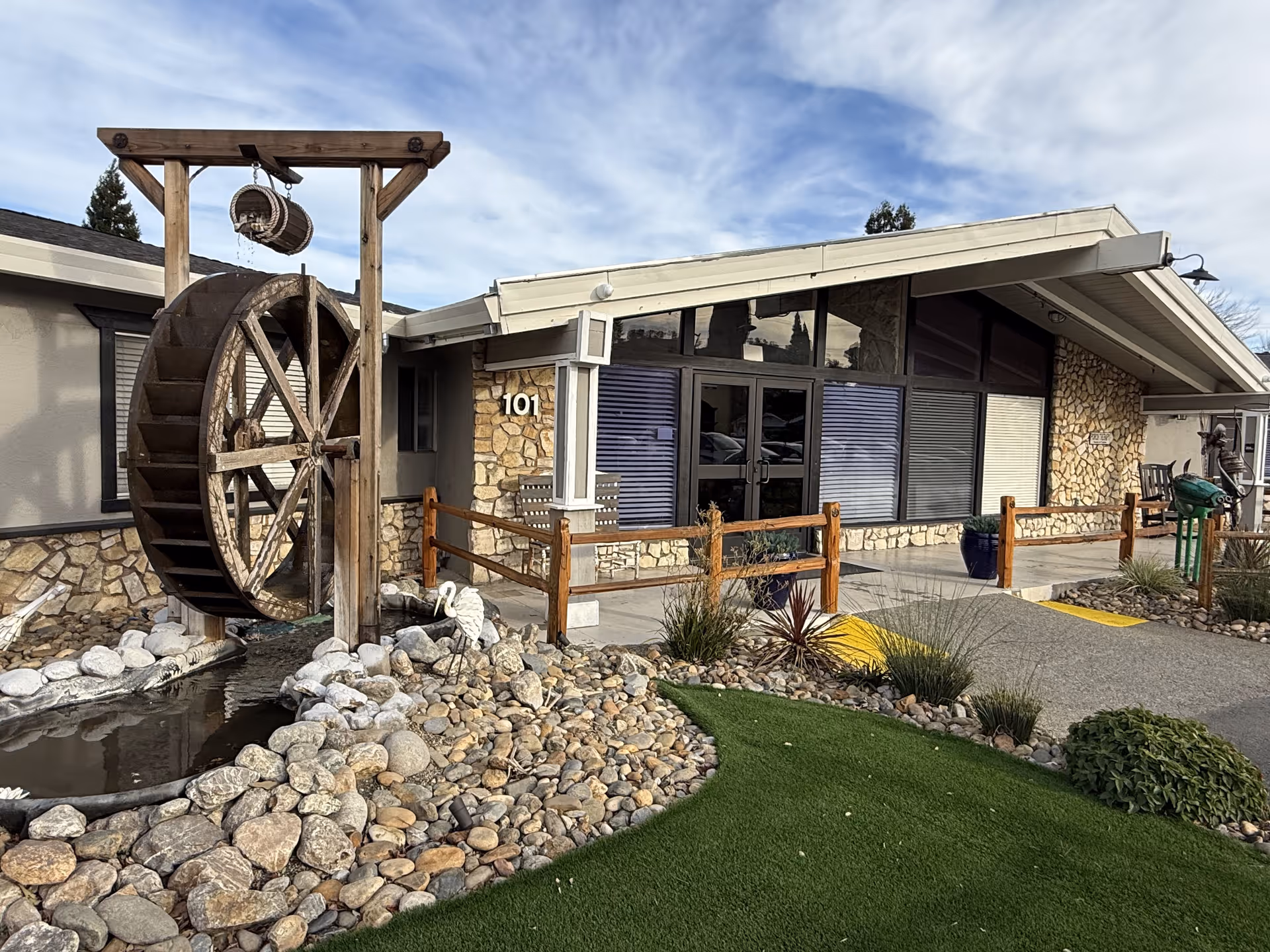 Stone-faced facility entrance with a large wooden waterwheel, rock garden and pathway leading to glass double doors.