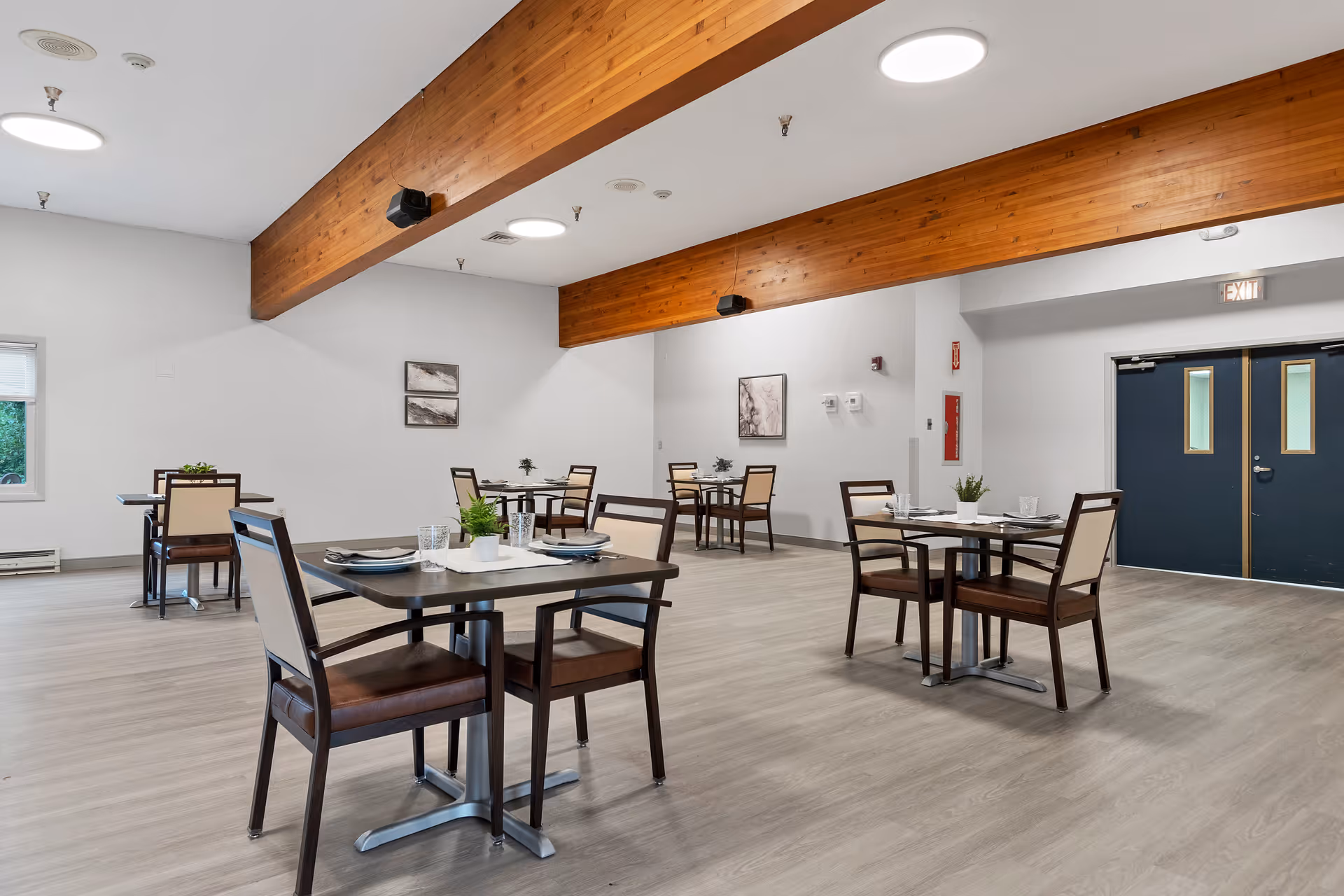 A spacious dining room with several square tables, each set with plates, glasses, and napkins. The room features light wood flooring, white walls, and exposed wooden beams on the ceiling. There are a few framed artworks on the walls and a double door with windows at the far end.