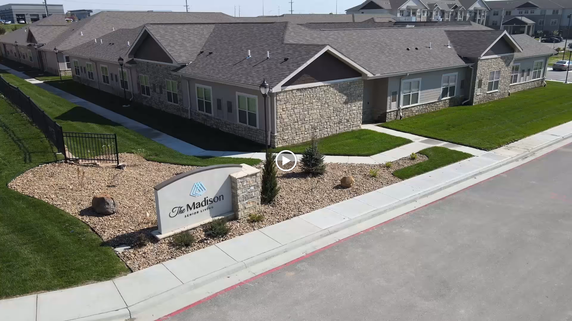 Exterior view of The Madison Senior Living facility showing a single-story building with stone and siding exterior, a landscaped area with rocks and small plants, a sidewalk, and a sign displaying the facility's name.