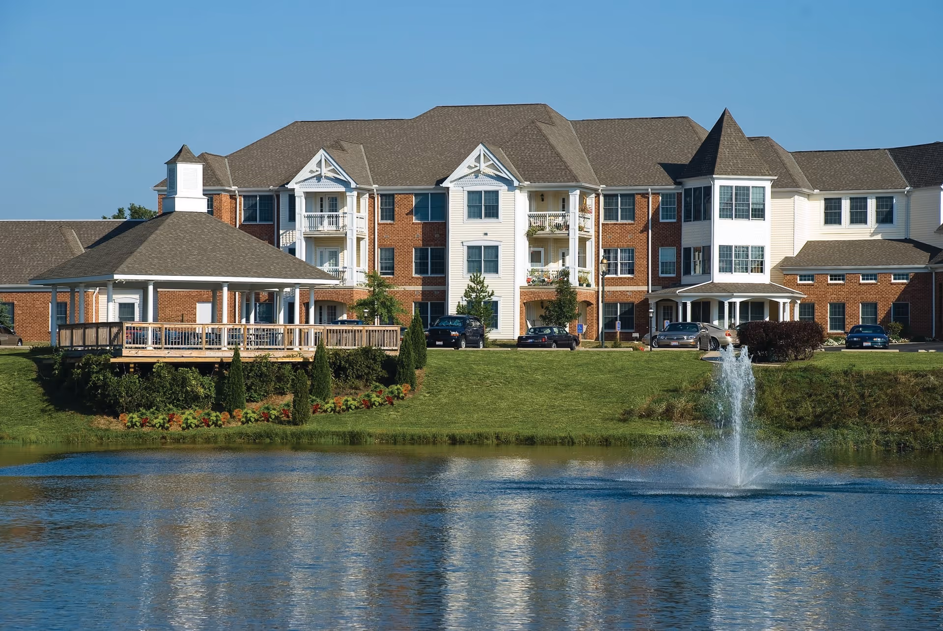 A large senior living facility building with a combination of brick and white siding, multiple balconies, and a gazebo on a grassy area next to a pond with a water fountain under a clear blue sky.