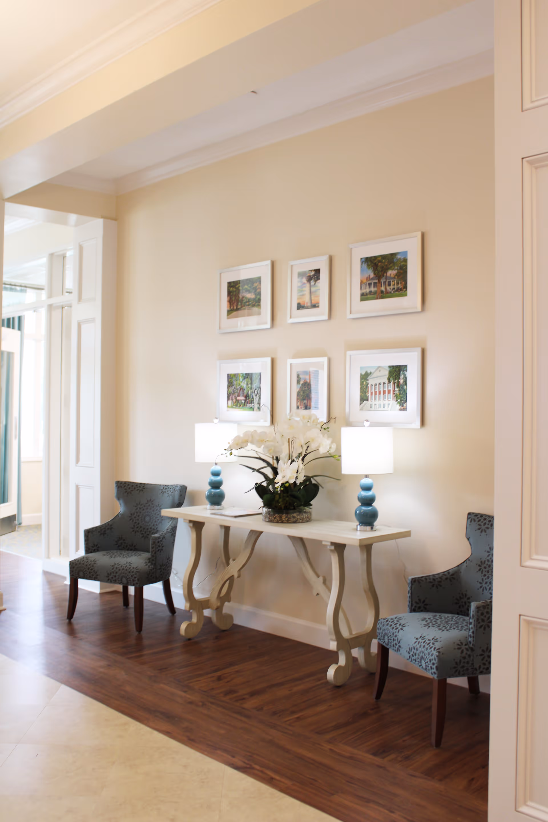 A cozy interior corner of The Homestead Assisted Living Community featuring a white decorative console table with a floral arrangement and two blue table lamps. Two patterned blue armchairs flank the table, and six framed pictures hang on the beige wall above. The floor is a combination of wood and tile, and natural light brightens the space.
