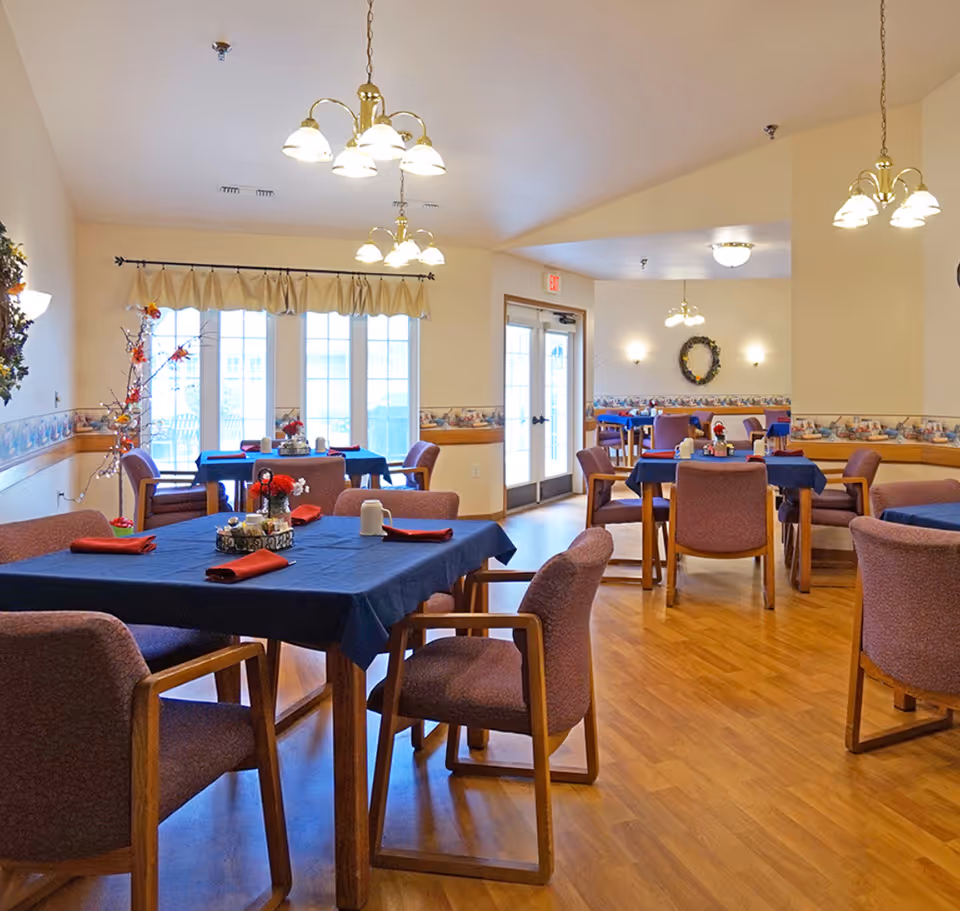 Bright communal dining room with multiple tables covered in blue tablecloths and chairs in an assisted living facility.