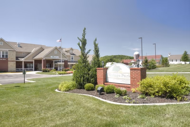 Exterior view of Vintage Park at Tonganoxie senior living facility with a manicured lawn, shrubs, and a brick sign in the foreground. The building has a beige and brick facade with a flagpole displaying an American flag near the entrance. The sky is clear and blue.