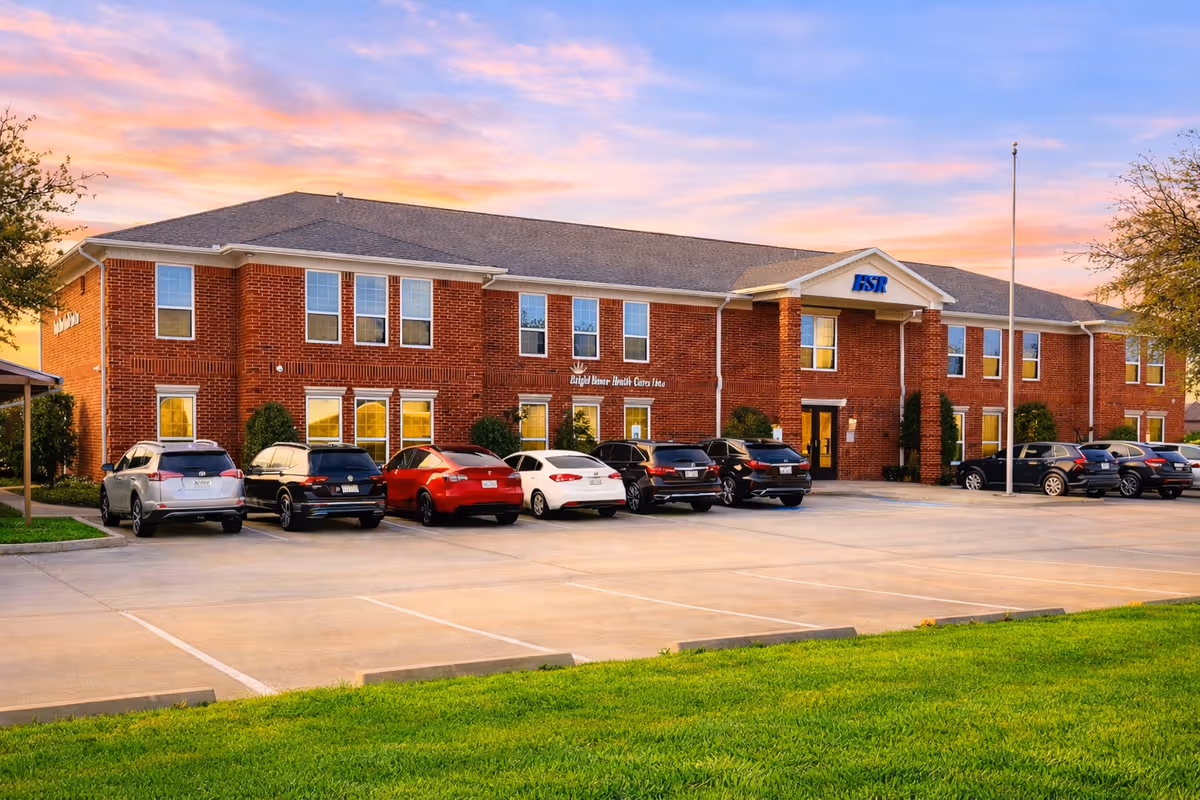 Brick senior living facility with a parking lot and parked cars at sunset.