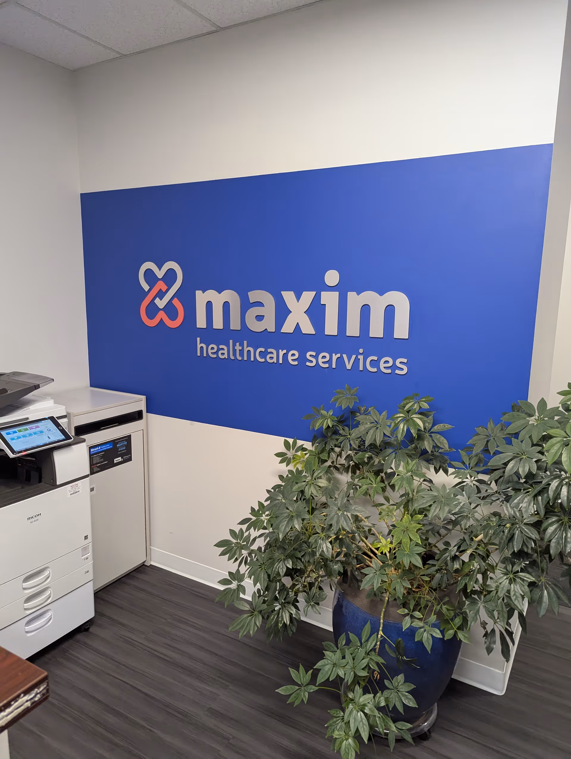 There is a blue Maxim Healthcare Services sign on the wall with a printer and plant.