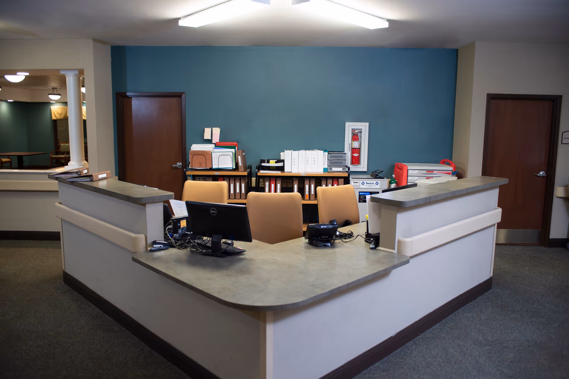 Reception desk area with computer monitors, chairs, and office supplies in front of a teal accent wall.