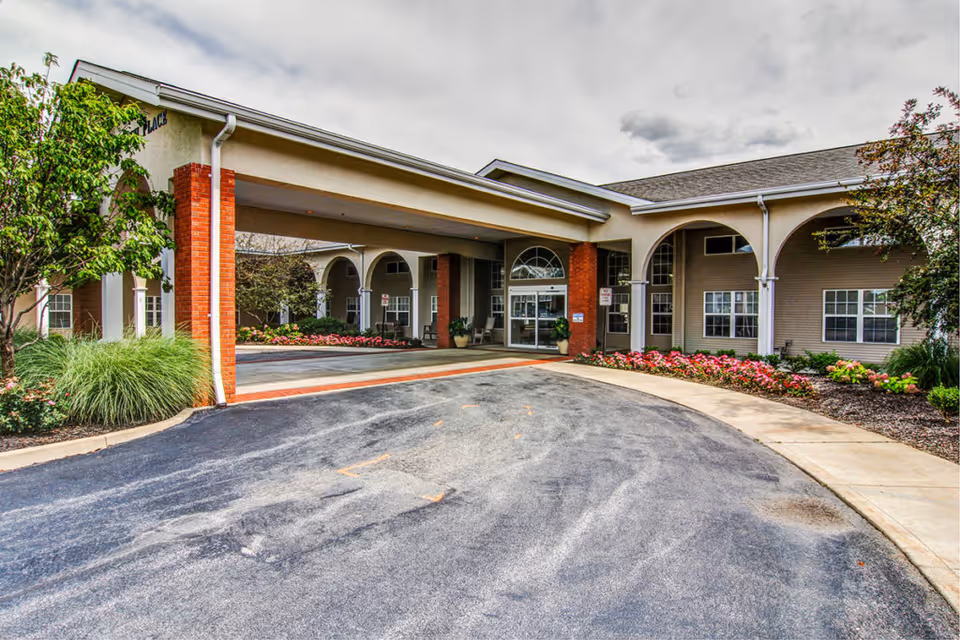 Front entrance of a senior living building with a covered porte-cochere, brick columns, driveway, and landscaped flower beds.