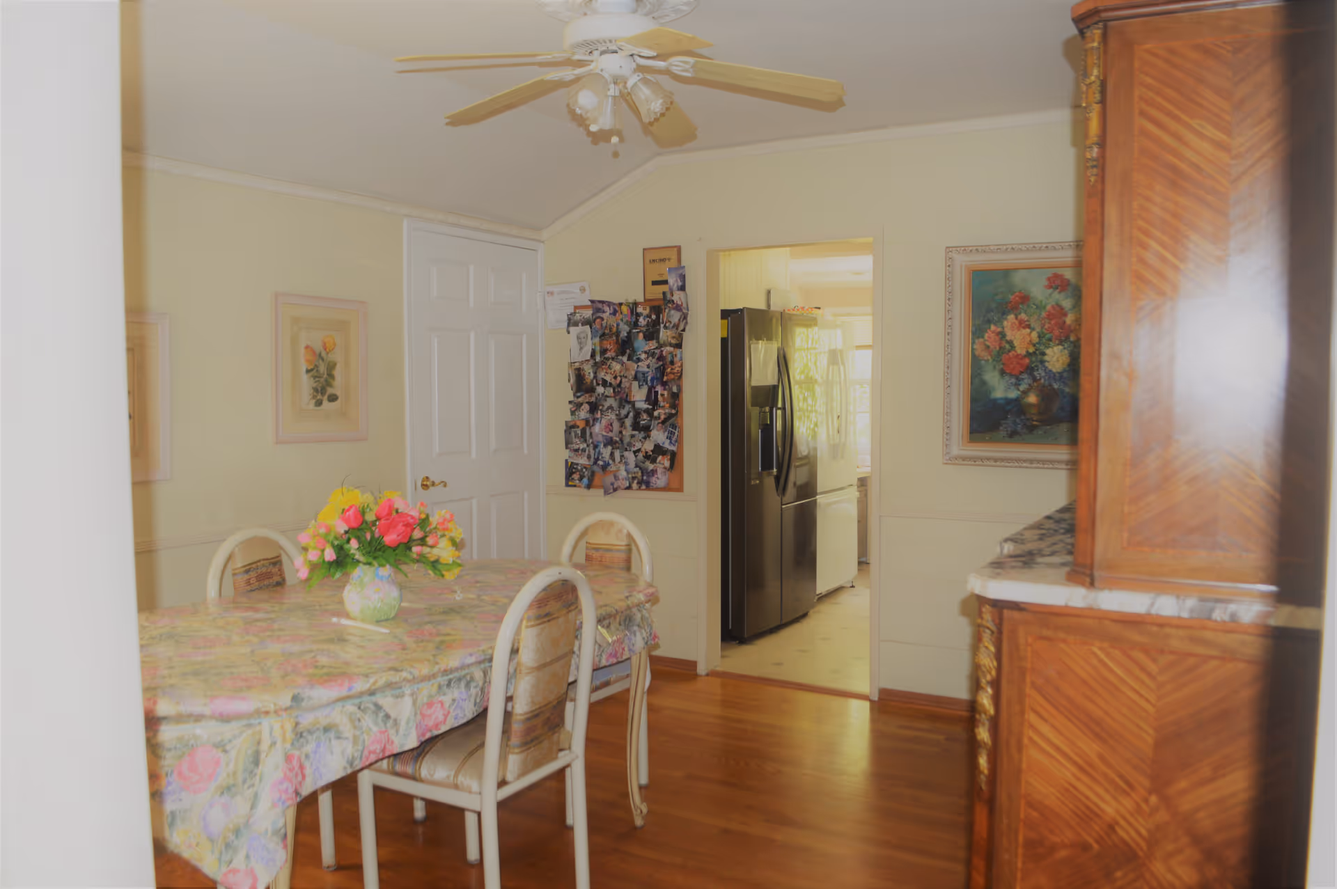 Dining room with a floral-covered table and chairs, a vase of flowers, ceiling fan, and a doorway opening to a kitchen with a refrigerator.