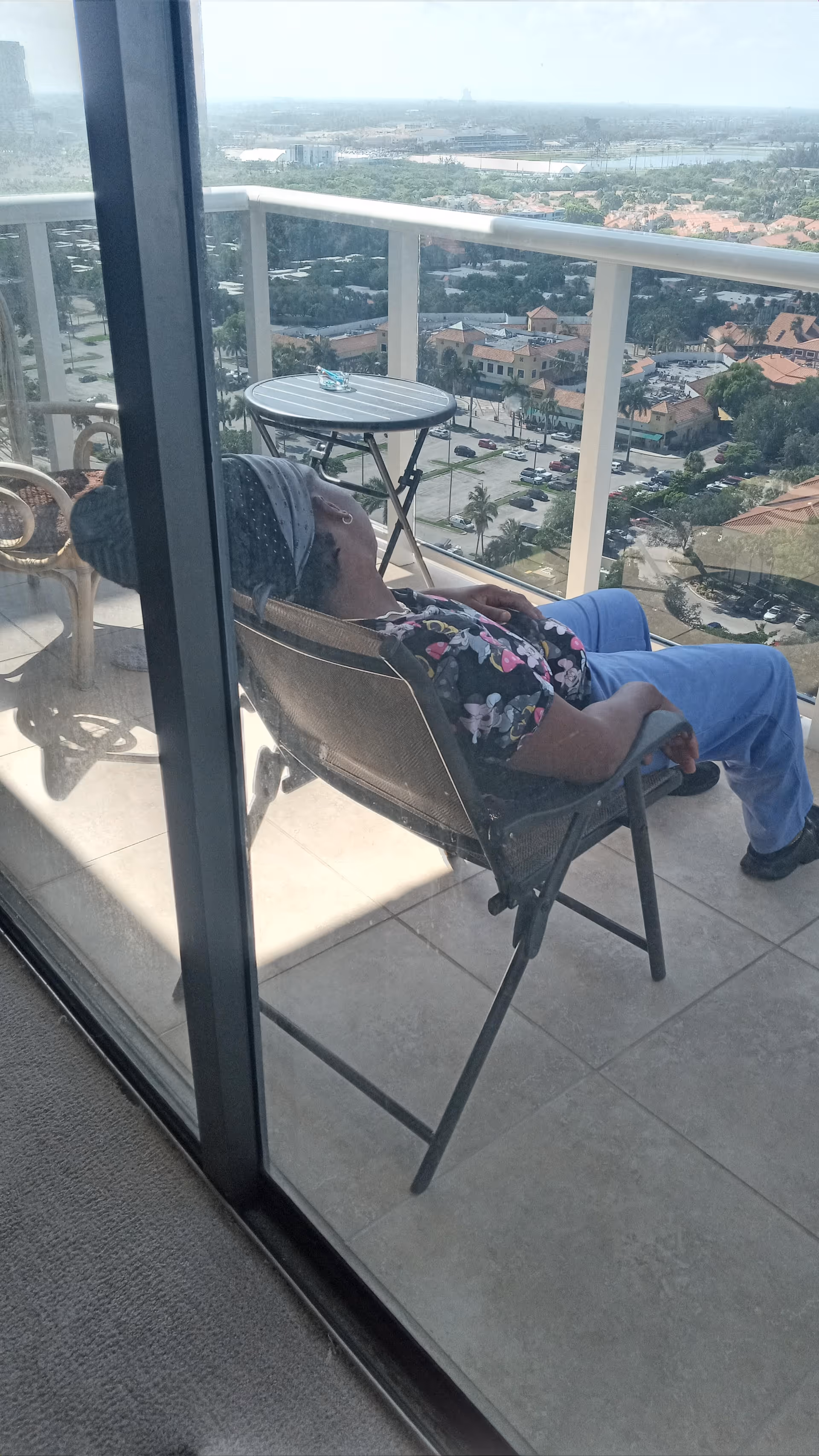 An elderly resident lounges on a balcony chair with a cityscape view.