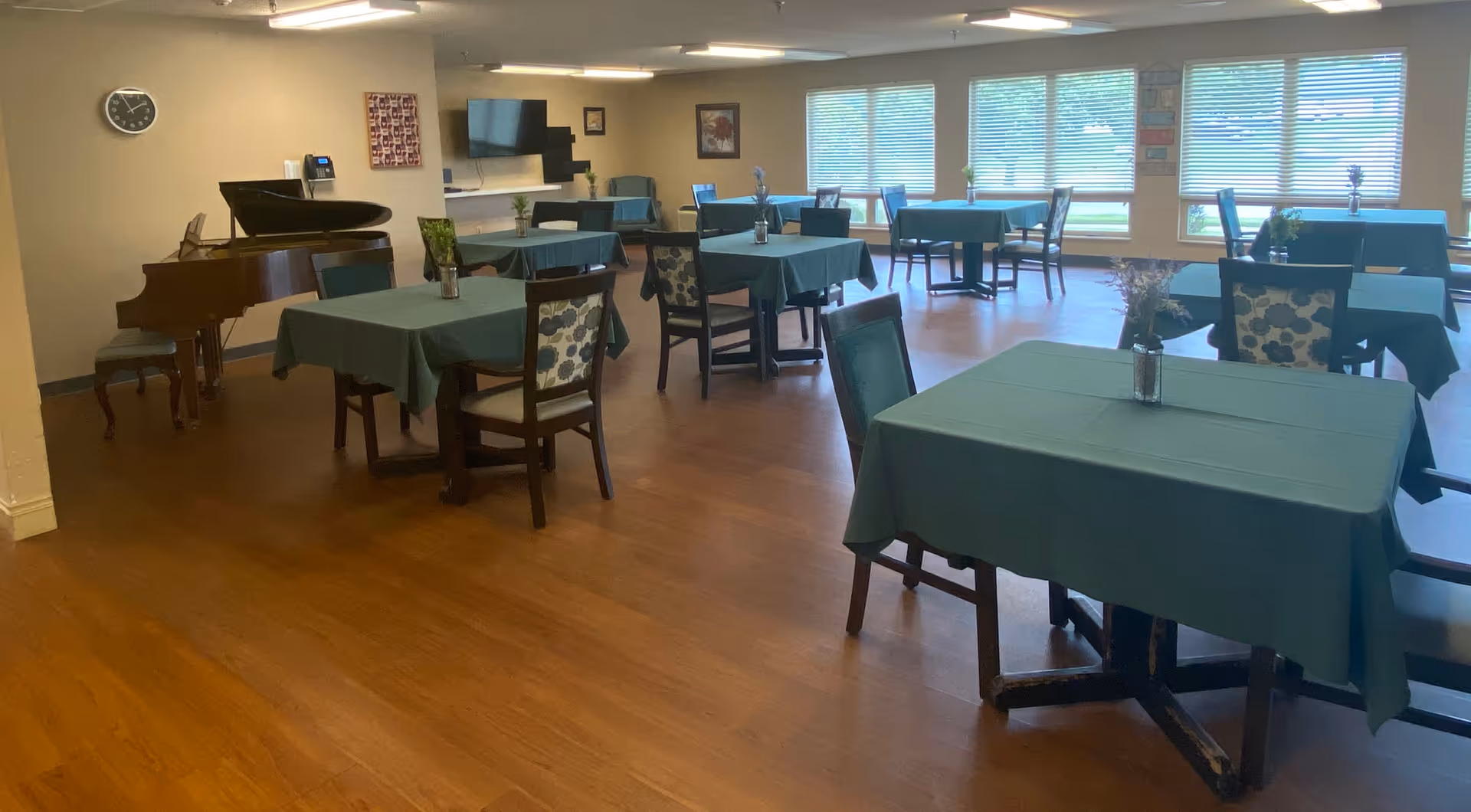 A spacious dining room with several tables covered in green tablecloths, each with a small vase of flowers. Chairs with patterned upholstery surround the tables. A grand piano is positioned against one wall, and a wall clock and a flat-screen TV are mounted on the walls. Large windows with blinds allow natural light to fill the room.