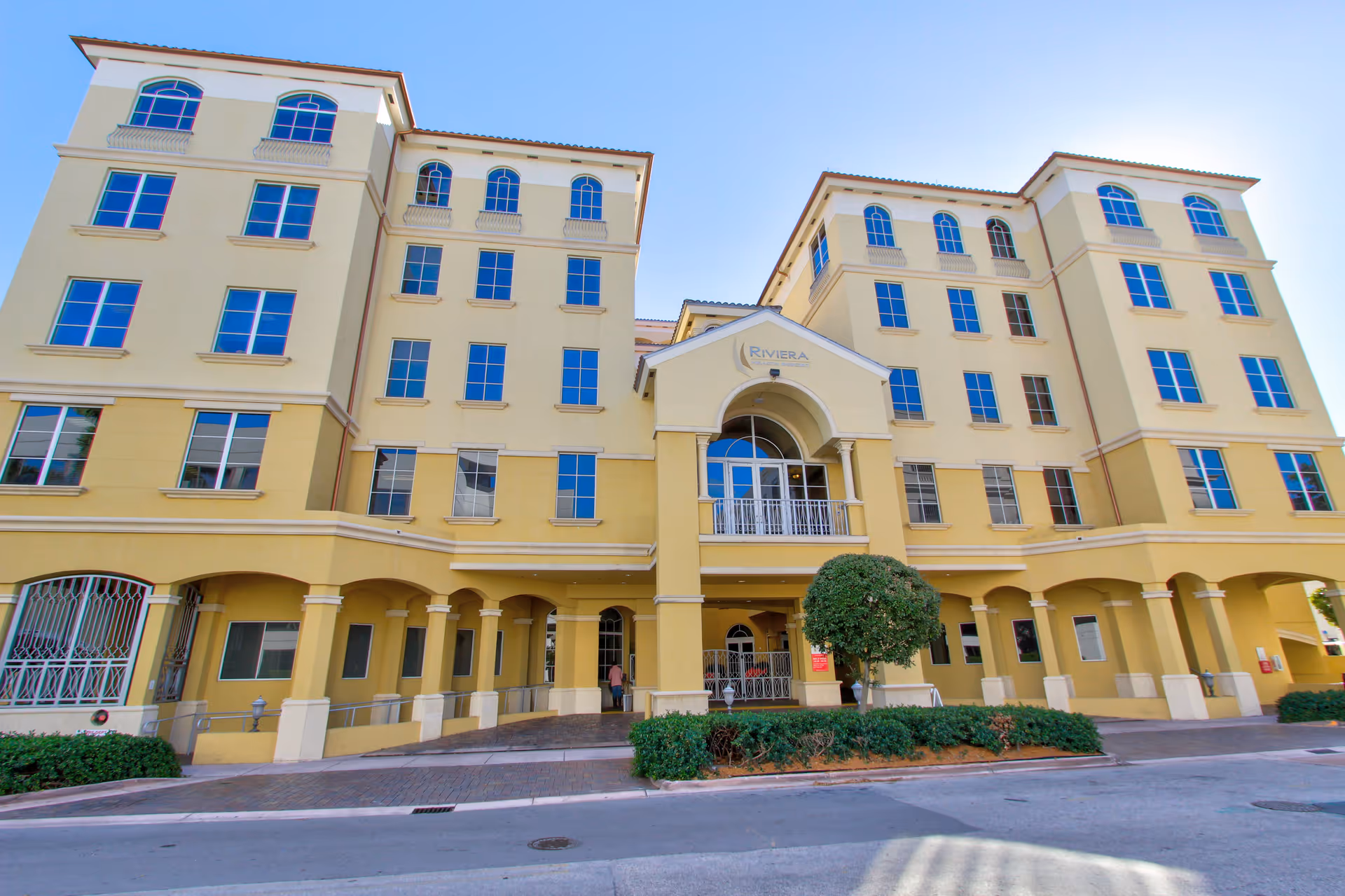 Front exterior view of a large, multi-story yellow building with arched windows and a central entrance labeled 'Riviera'. The building has a Mediterranean architectural style with a tiled roof and columns supporting a covered walkway.