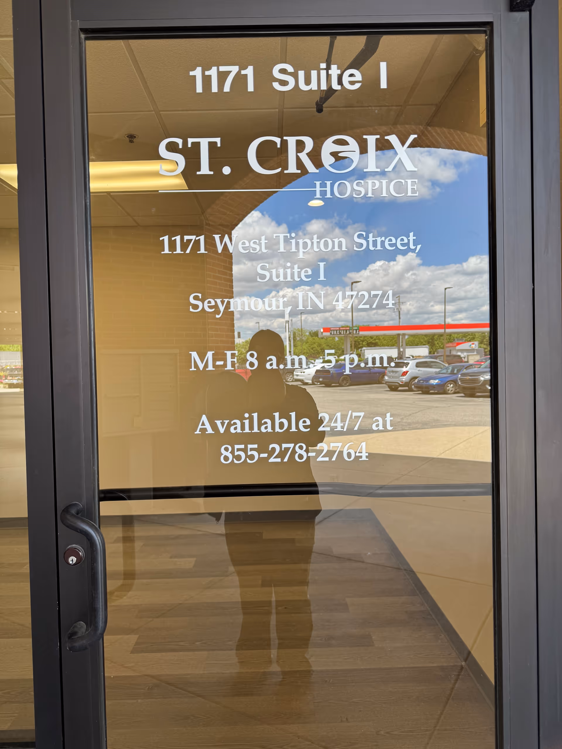 Glass door displays St. Croix Hospice sign with address and hours, reflecting parking lot outside.