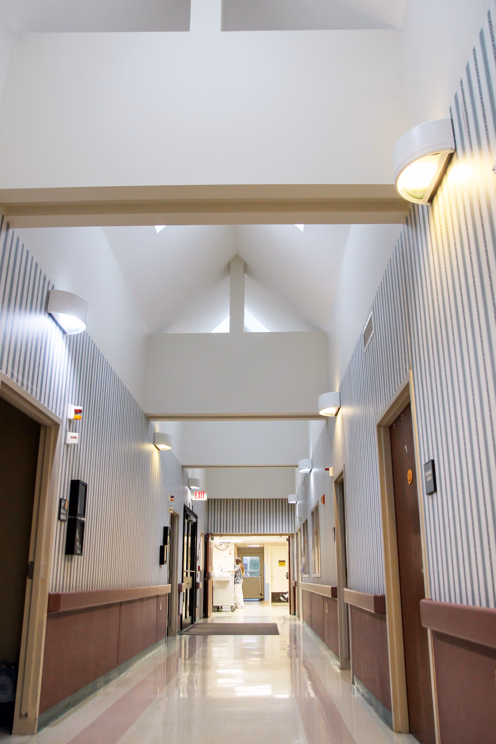 Bright vaulted interior hallway of a care facility with striped walls, wall sconces, doors on both sides and a person visible at the far end.