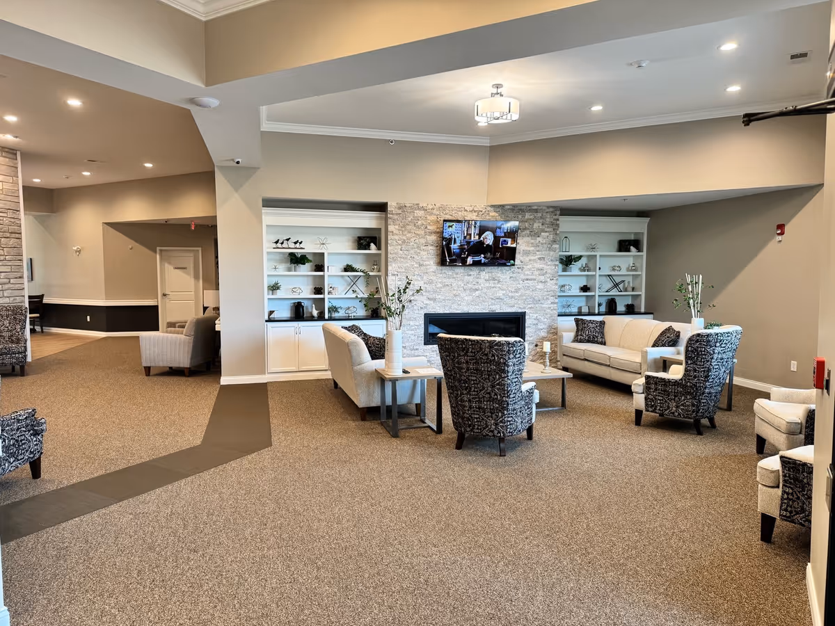 A spacious and well-lit living room area in an assisted living facility featuring a stone fireplace with a mounted flat-screen TV above it. The room has beige walls and carpeted floors with a dark patterned pathway. There are multiple seating options including two beige sofas, two patterned armchairs, and additional chairs arranged around coffee tables. Built-in white shelves flank the fireplace, decorated with plants and ornaments. Recessed lighting and a ceiling light fixture illuminate the room.