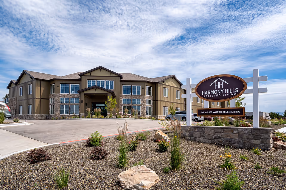Exterior view of a two-story assisted living facility building with large windows and a stone and wood facade under a partly cloudy sky. In the foreground, there is a landscaped area with rocks, plants, and a sign that reads 'Harmony Hills Assisted Living - Live a Life Worth Celebrating.'