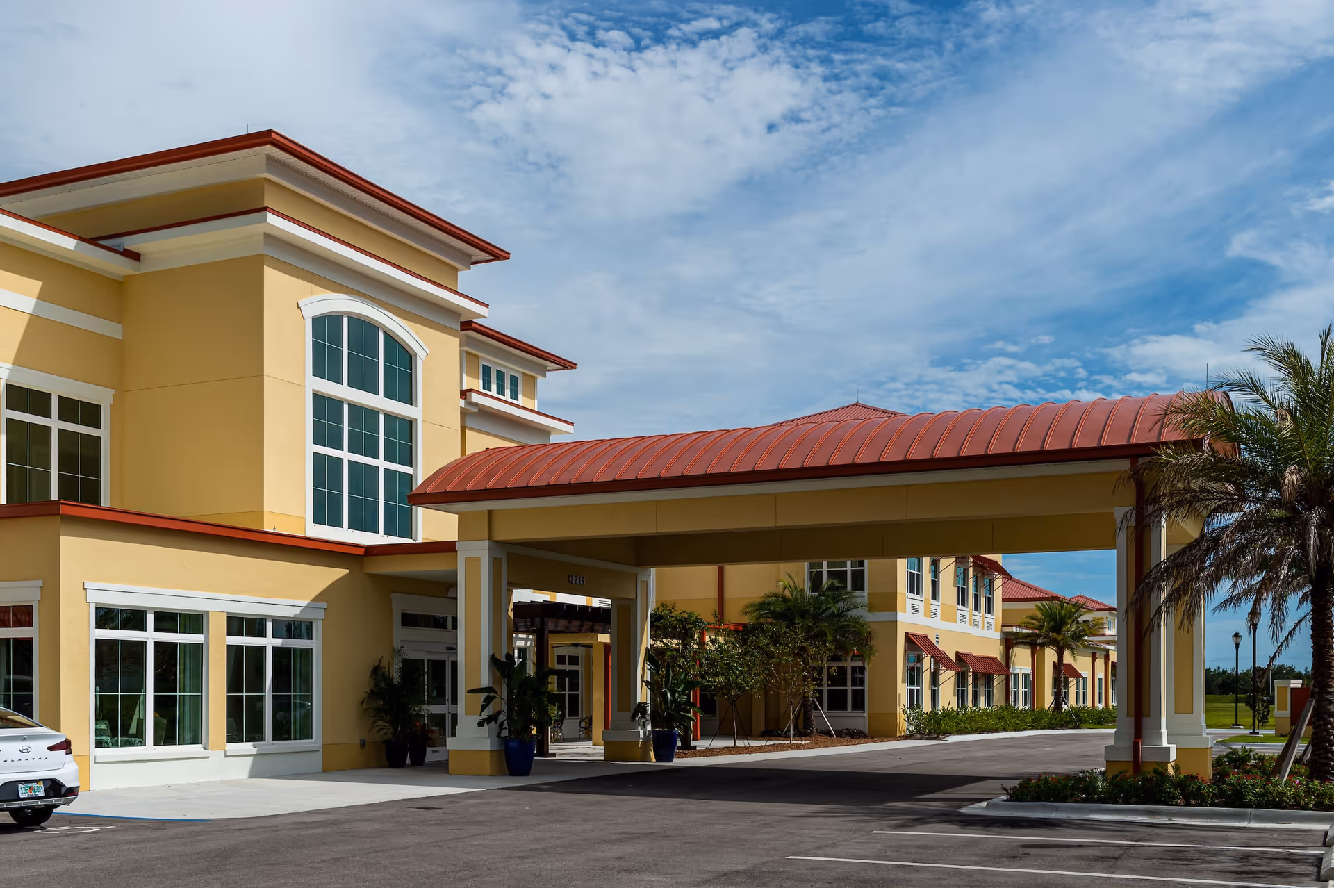 Exterior view of a yellow senior living facility building with large windows and a red roof canopy over the entrance. There are palm trees and plants around the entrance area under a partly cloudy sky.
