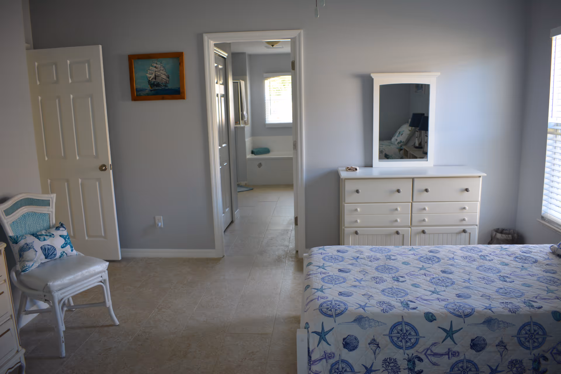 Bright bedroom with blue walls, white furniture, patterned bedspread, and adjacent bathroom doorway.