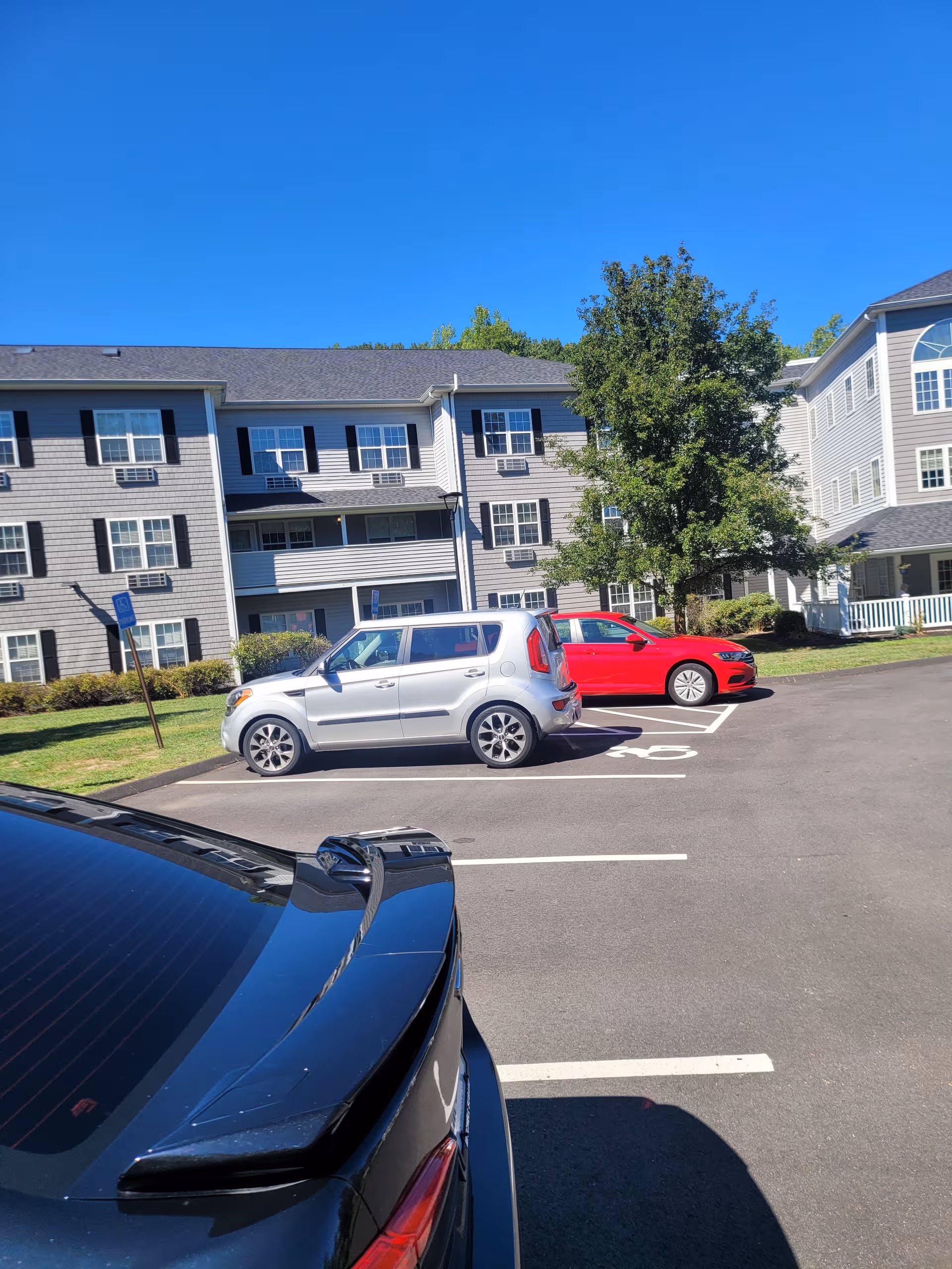 Three-story gray senior living building with white trim, parked cars, and a blue sky.