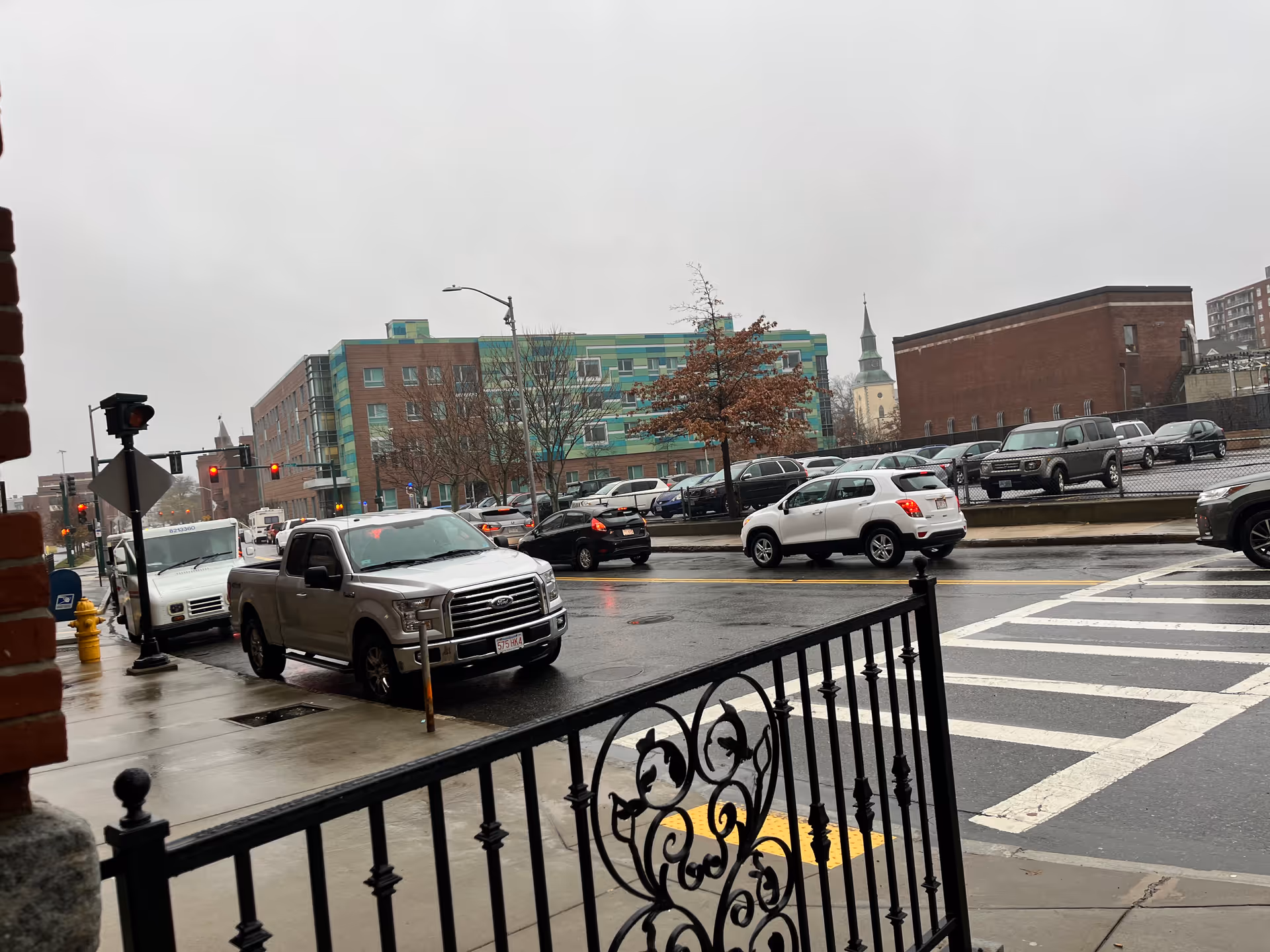 Rainy city street with cars, crosswalk, and decorative railing in foreground.