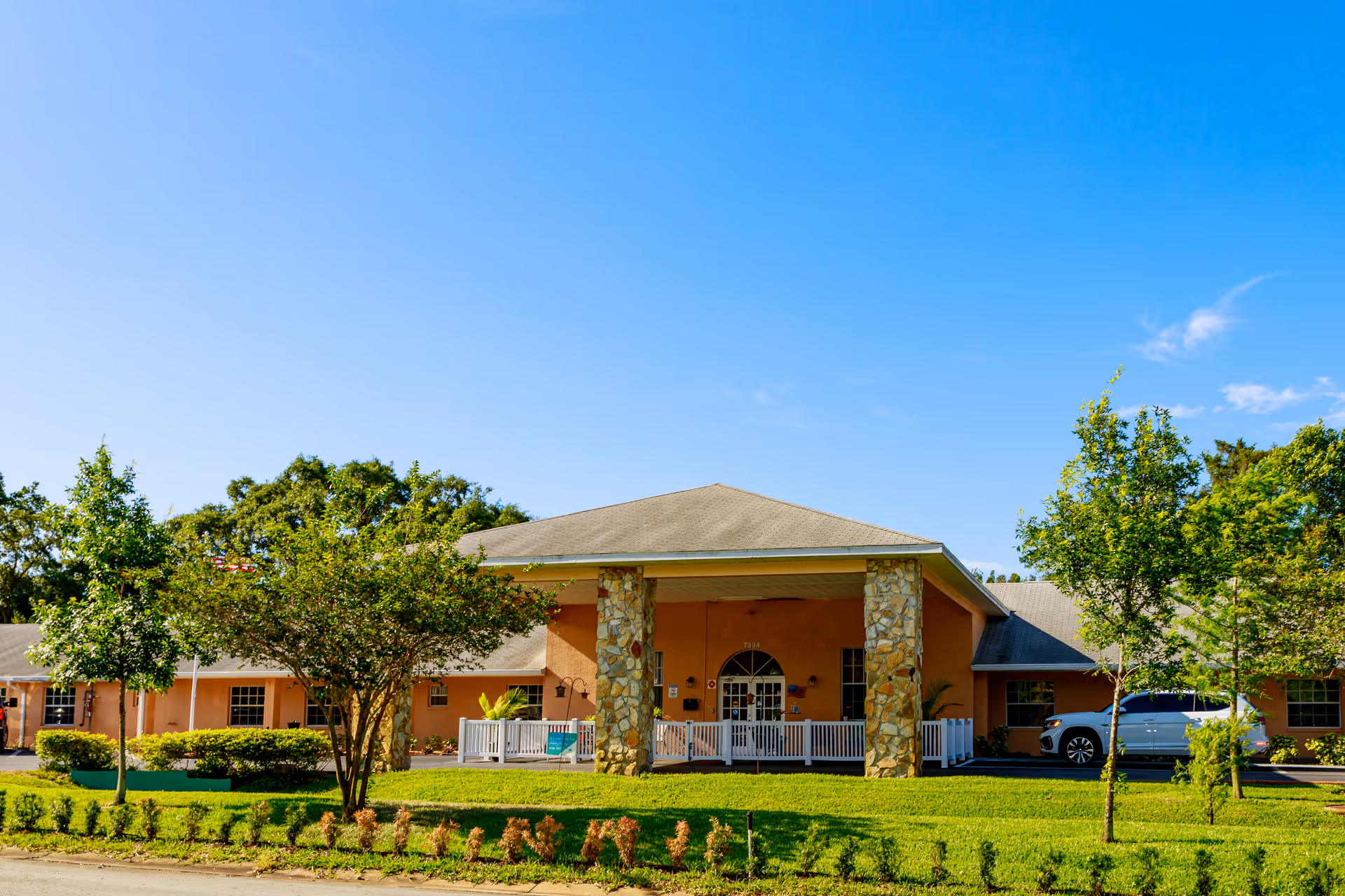 Sunny exterior view of Jennifer Gardens senior living facility with entrance, porch, and manicured grounds.