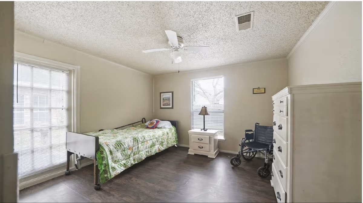 A simple bedroom with a single bed, nightstand and lamp, dresser, ceiling fan and a wheelchair by the window.