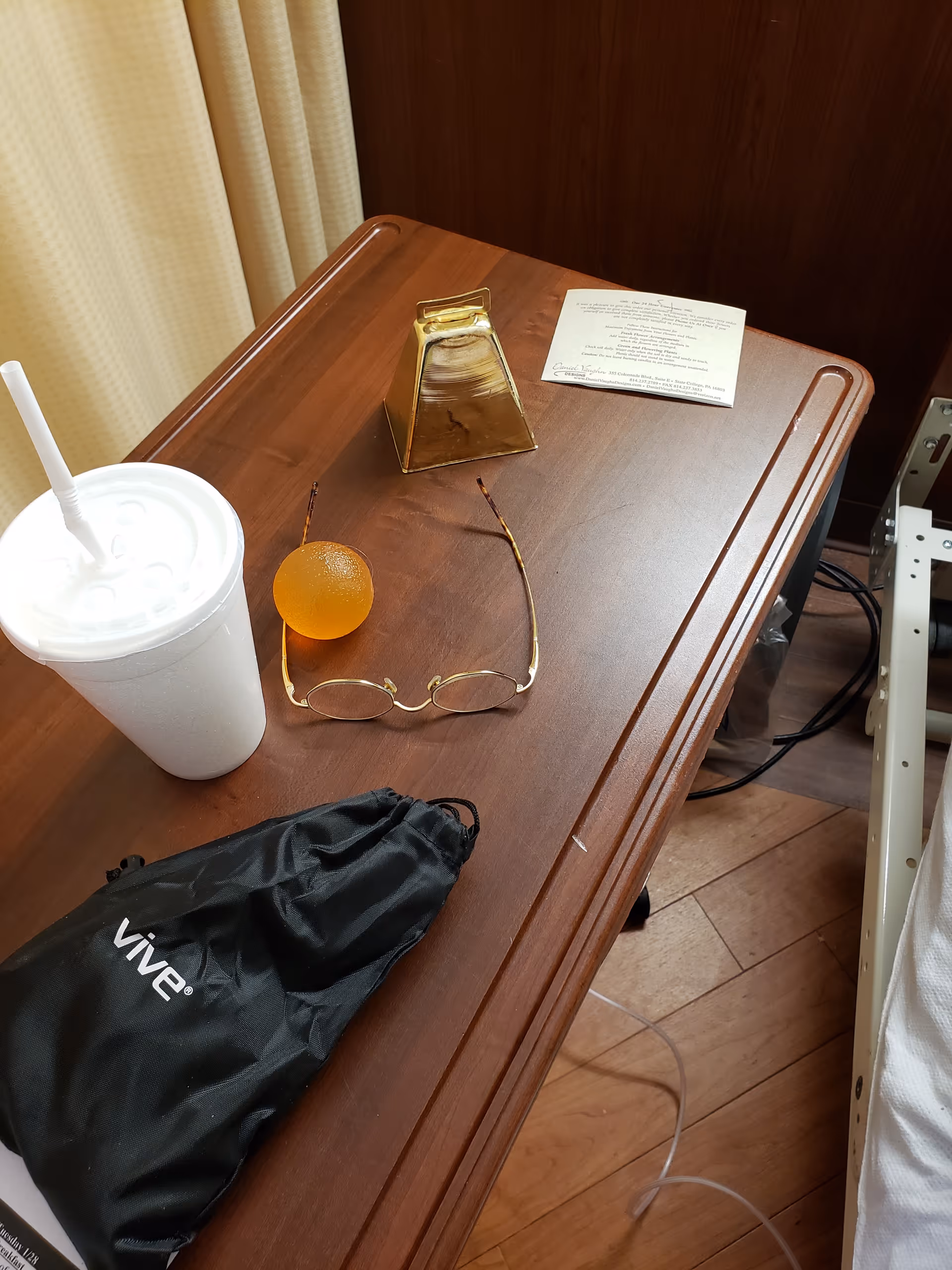 Wooden bedside table with cup, glasses, orange ball, bell, and documents in a patient room.