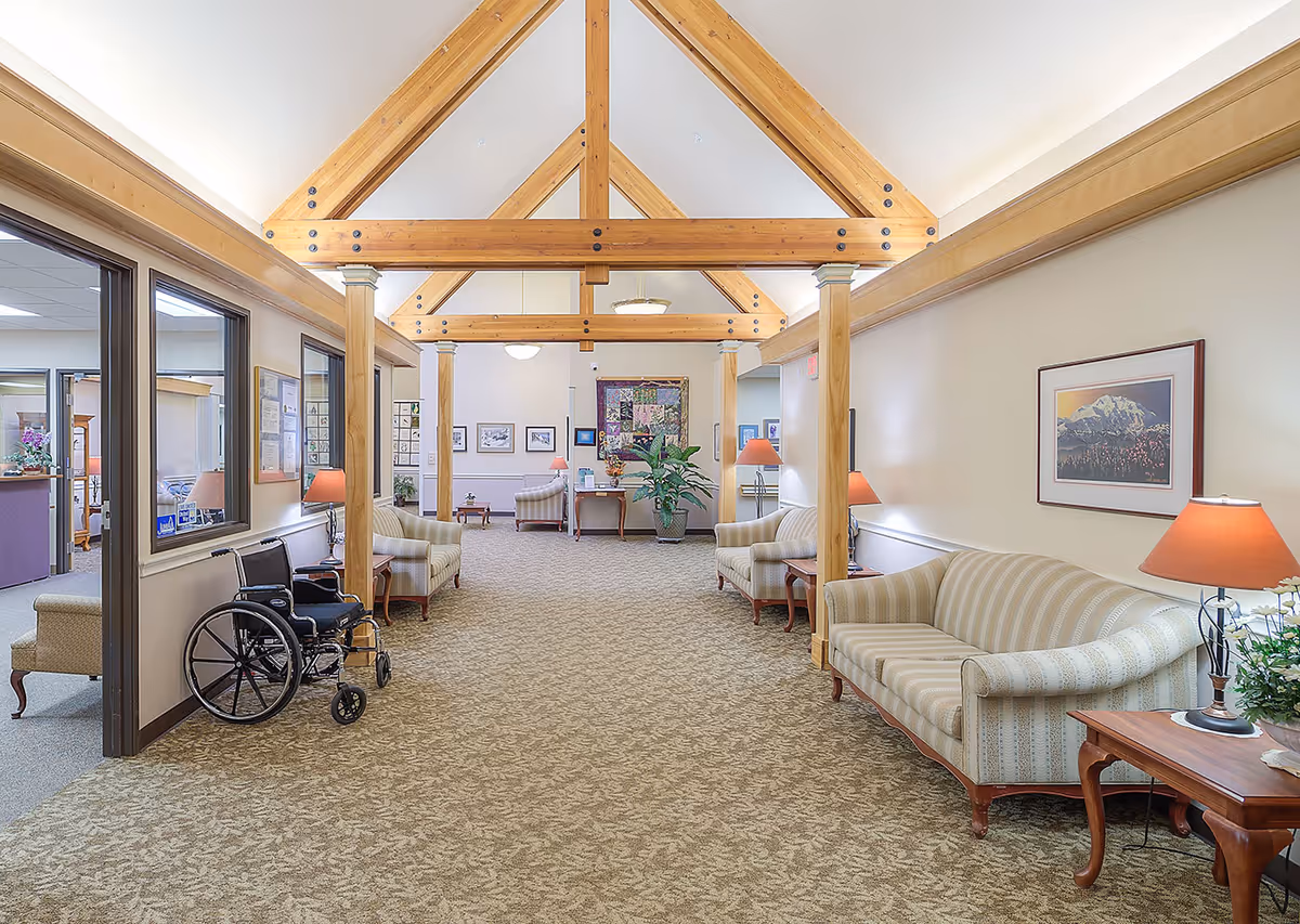 Senior living lounge with vaulted wooden beams, striped sofas, side tables with lamps, plants and a wheelchair near the entrance.