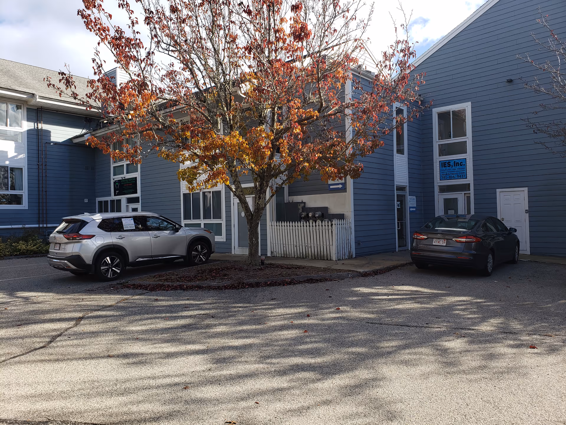 Blue senior living building exterior with parked cars, a tree, and a white fence.