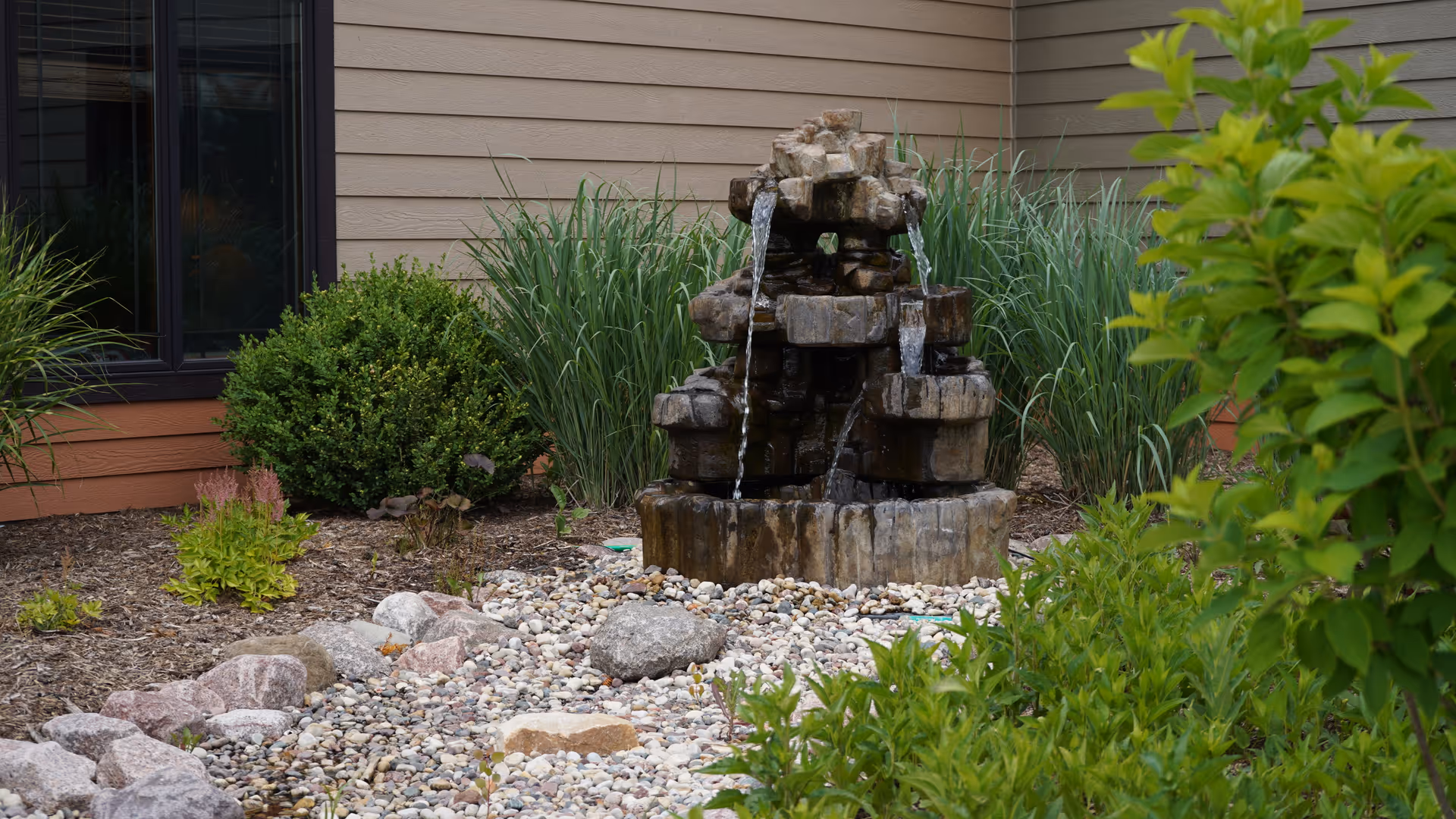Outdoor garden area with a multi-tiered stone water fountain surrounded by small rocks, green bushes, and tall grass plants, next to the exterior wall of a building with beige siding and a window.