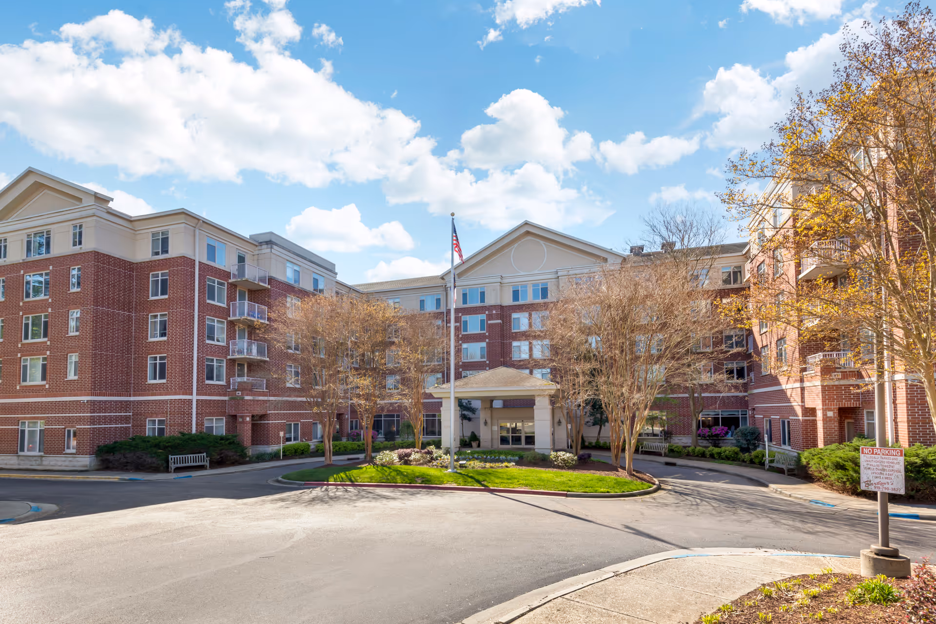 Brick senior living facility with central entrance, flagpole, and circular driveway under blue sky.