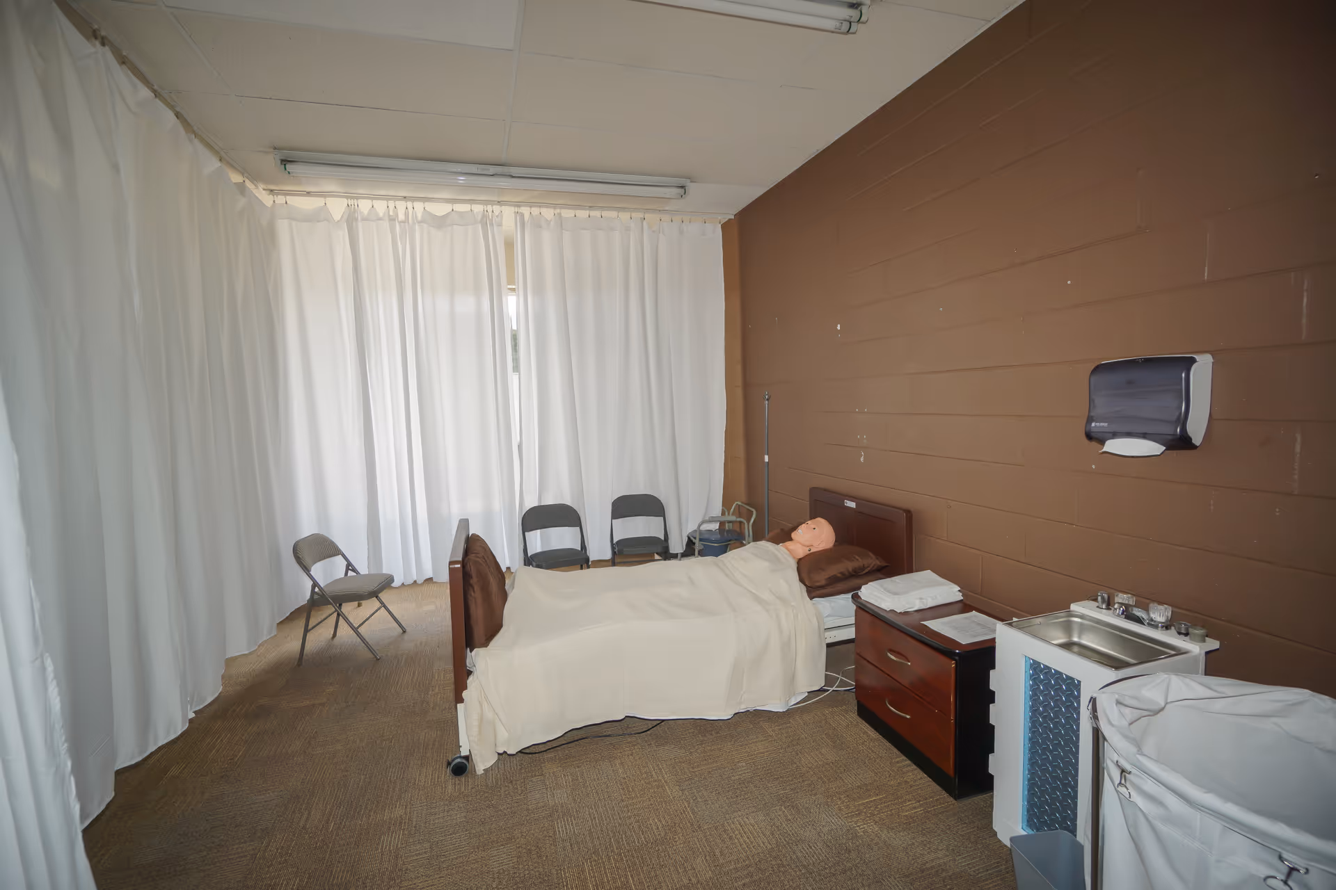 A hospital-style bedroom with a bed, white curtains, a sink, and chairs along the wall.
