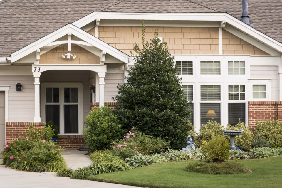 Front exterior view of a single-story residential building with beige siding, brick accents, and a small covered porch. The porch has white columns and the number 73 displayed. There are several bushes, flowering plants, a small statue, and a birdbath in the landscaped front yard.
