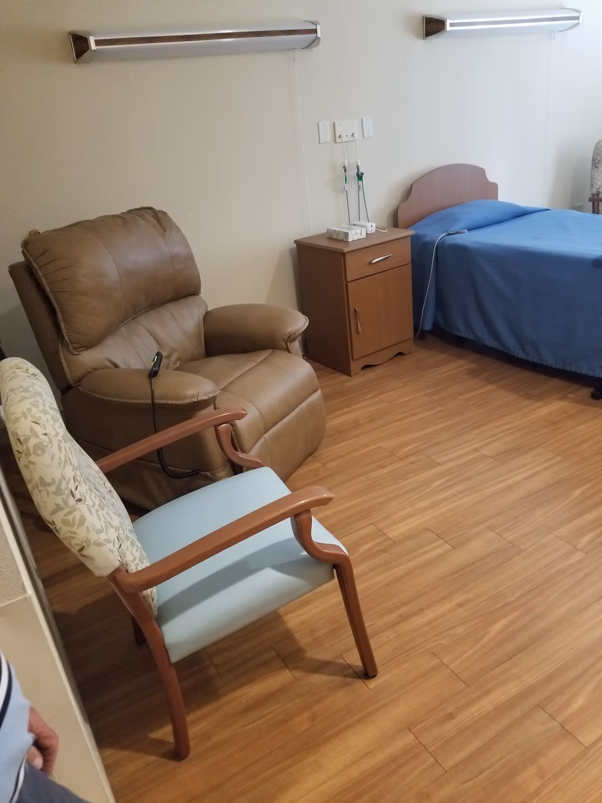 Interior room in a senior living facility with bed, nightstand, and chairs.