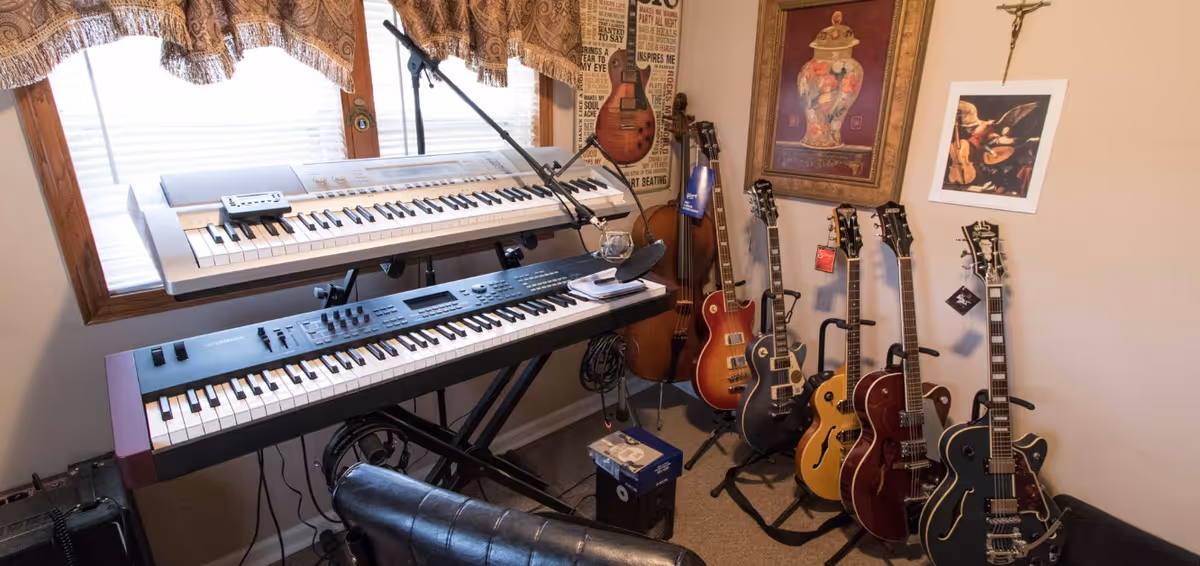 A music room with two electronic keyboards on stands in front of a window with blinds and a valance. Several guitars are displayed on stands against the wall, along with framed artwork and a microphone stand nearby. A black leather chair is partially visible in the foreground.